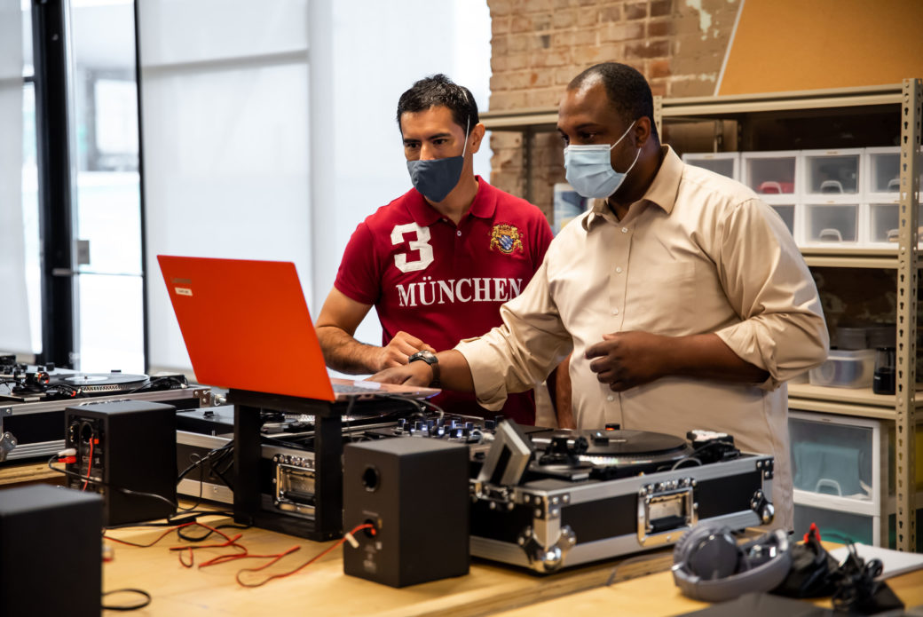 Two men stand behind a DJ turn table while looking into a red laptop computer