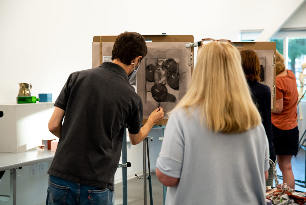 Instructor gives a demonstration in front of student standing in front of a drawing easel.