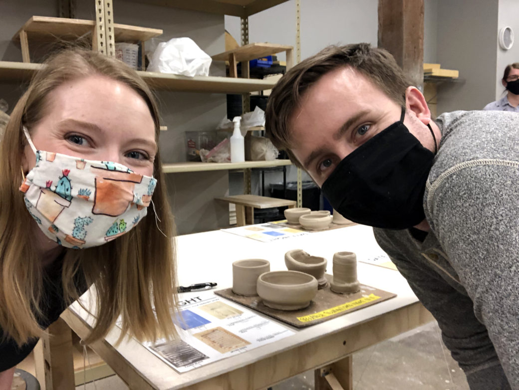 Two masked people looking at the camera with clay objects on the table between them