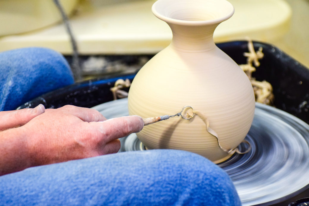 Close up image of two hands holding potters tools manipulating a vase thats spinning on a potter's wheel.