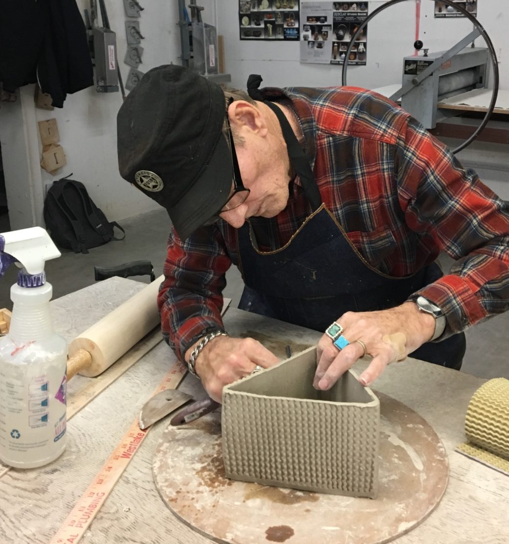 Person in a flannel shirt and black hat works on a triangular clay sculpture