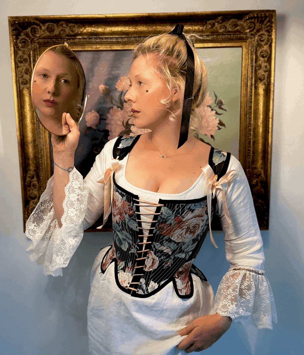 A person in a white shift and corset looks at their reflection in a handheld mirror