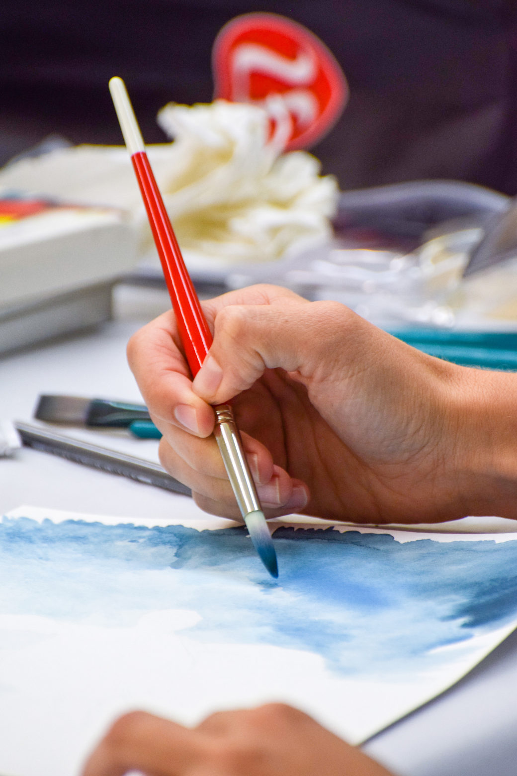 Image of a slender hand holding a paintbrush stroking a page with blue watercolors