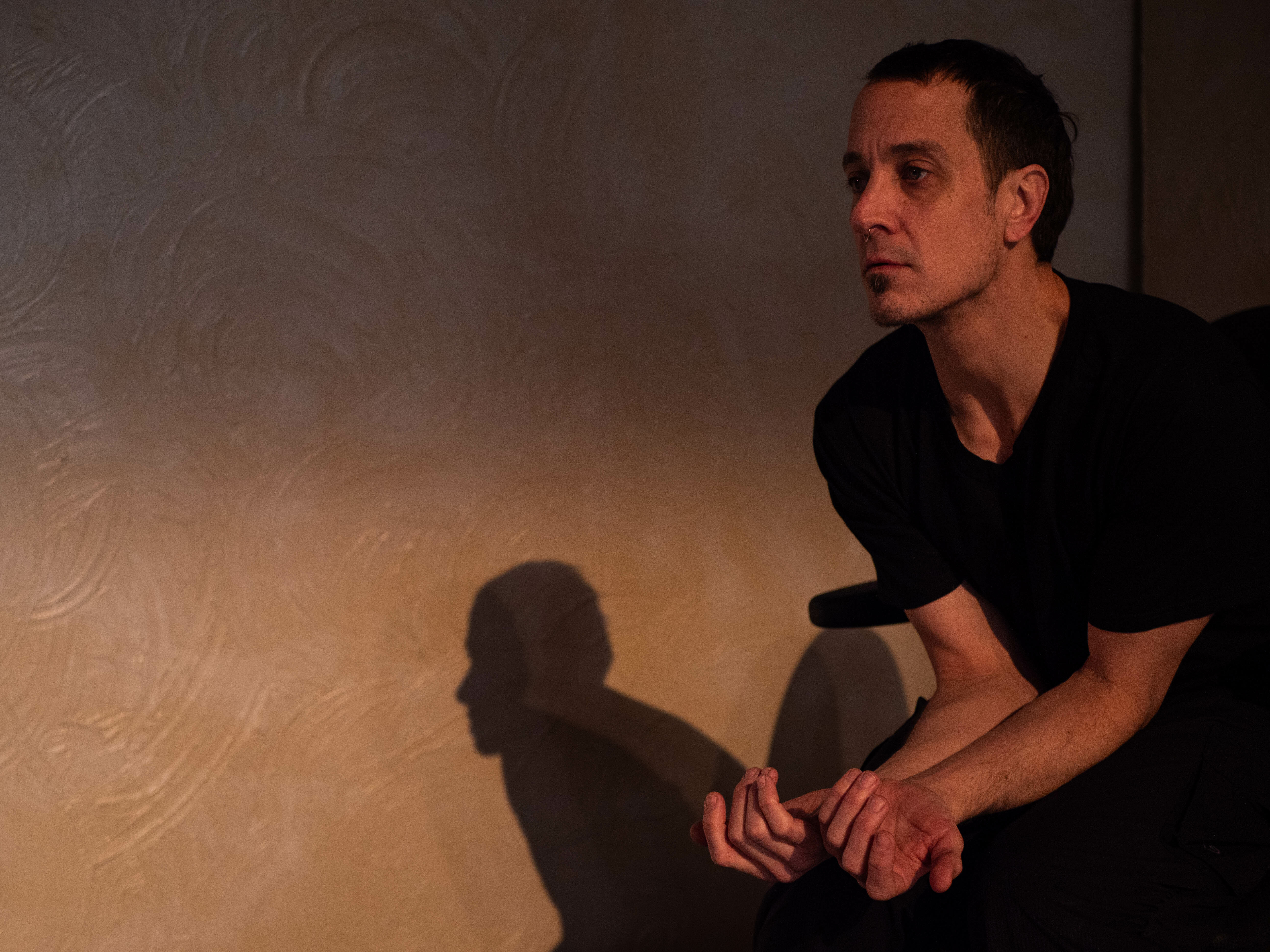 Lit by dark, glowing light, a dancer is hunched over, arms bent in front. They are dressed in a black shirt and their shadow shows behind them.