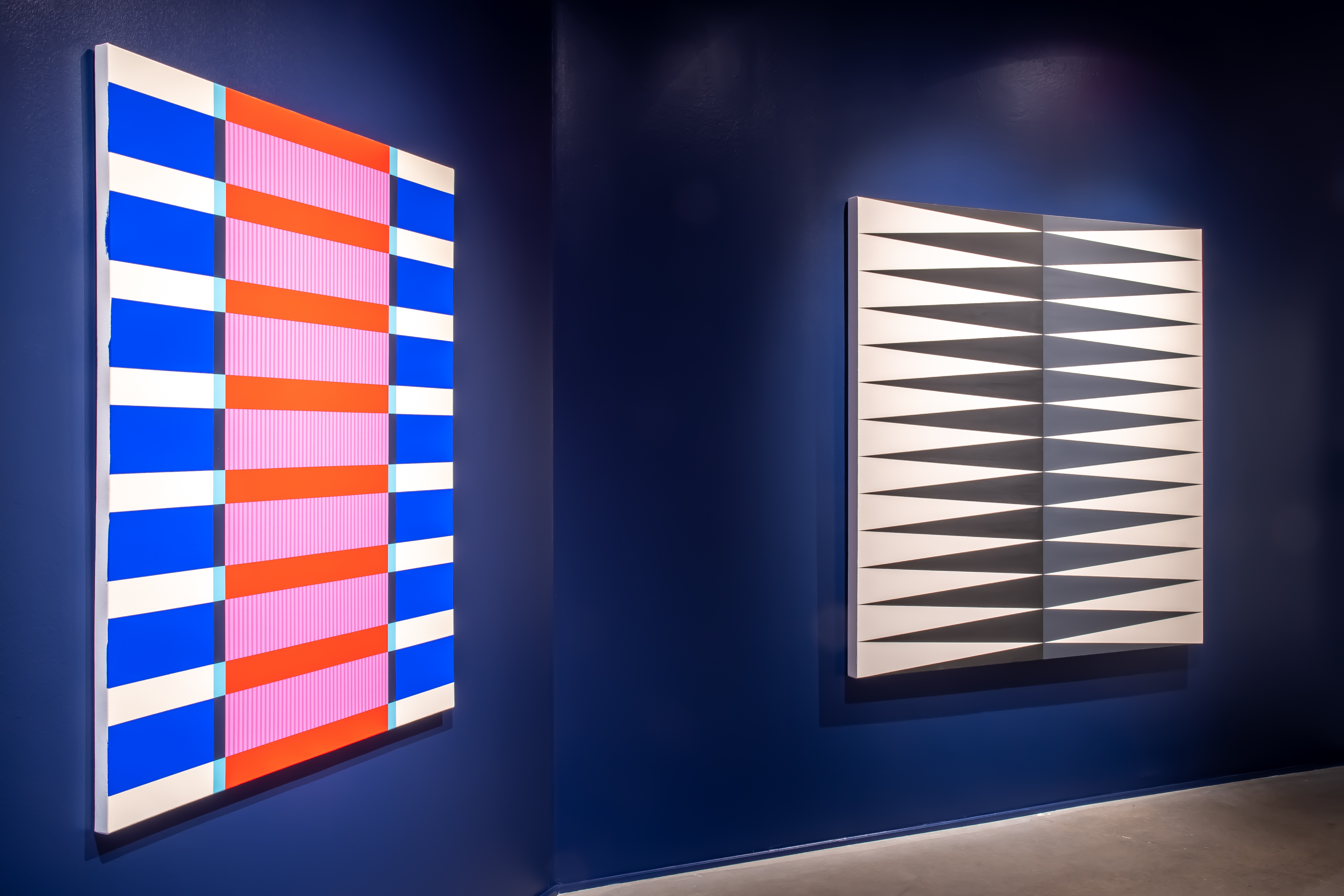 Two large paintings hang on a dark blue wall. On the left: Stark blue, white, red and pink lines create a pattern of three columns with alternating colors. Right: Two shades of gray pointed shapes in two columns create teeth-like imagery on a white canvas