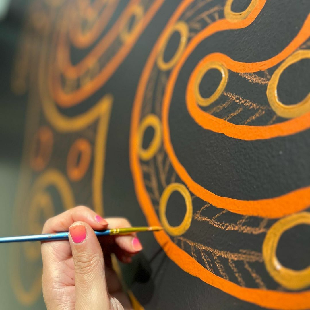 A close up image of a hand holding a blue-handled paintbrush painting an orange circle onto a black wall with circular orange patterns.