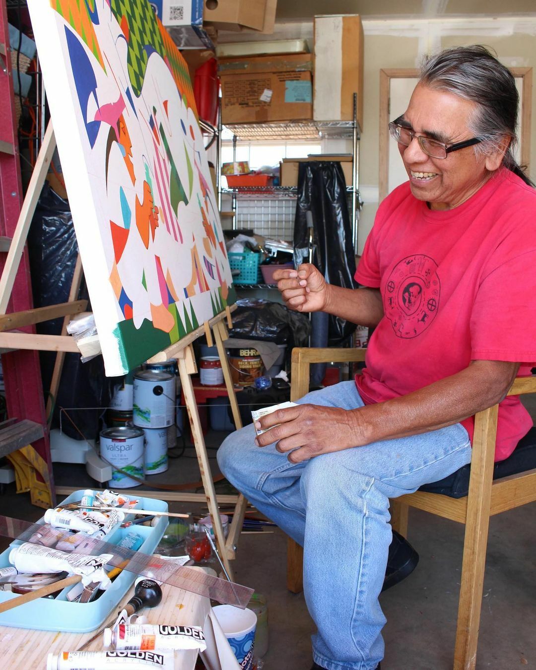 An Indigenous man in a red shirt, blue jeans and thick-framed glasses sits in front of a colorful, partially painted canvas on a wooden easel. The man is smiling and holding a paint brush.