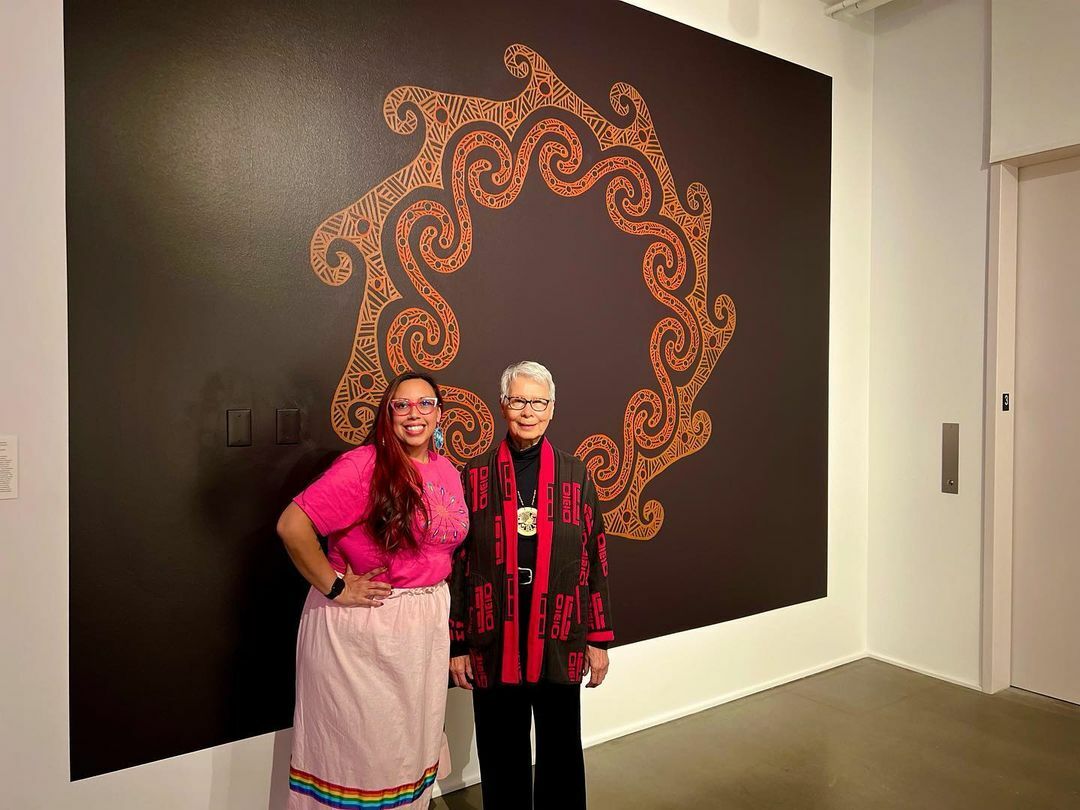 Two Indigenous women stand in front of a large, circular mural in orange pigment on a black background.