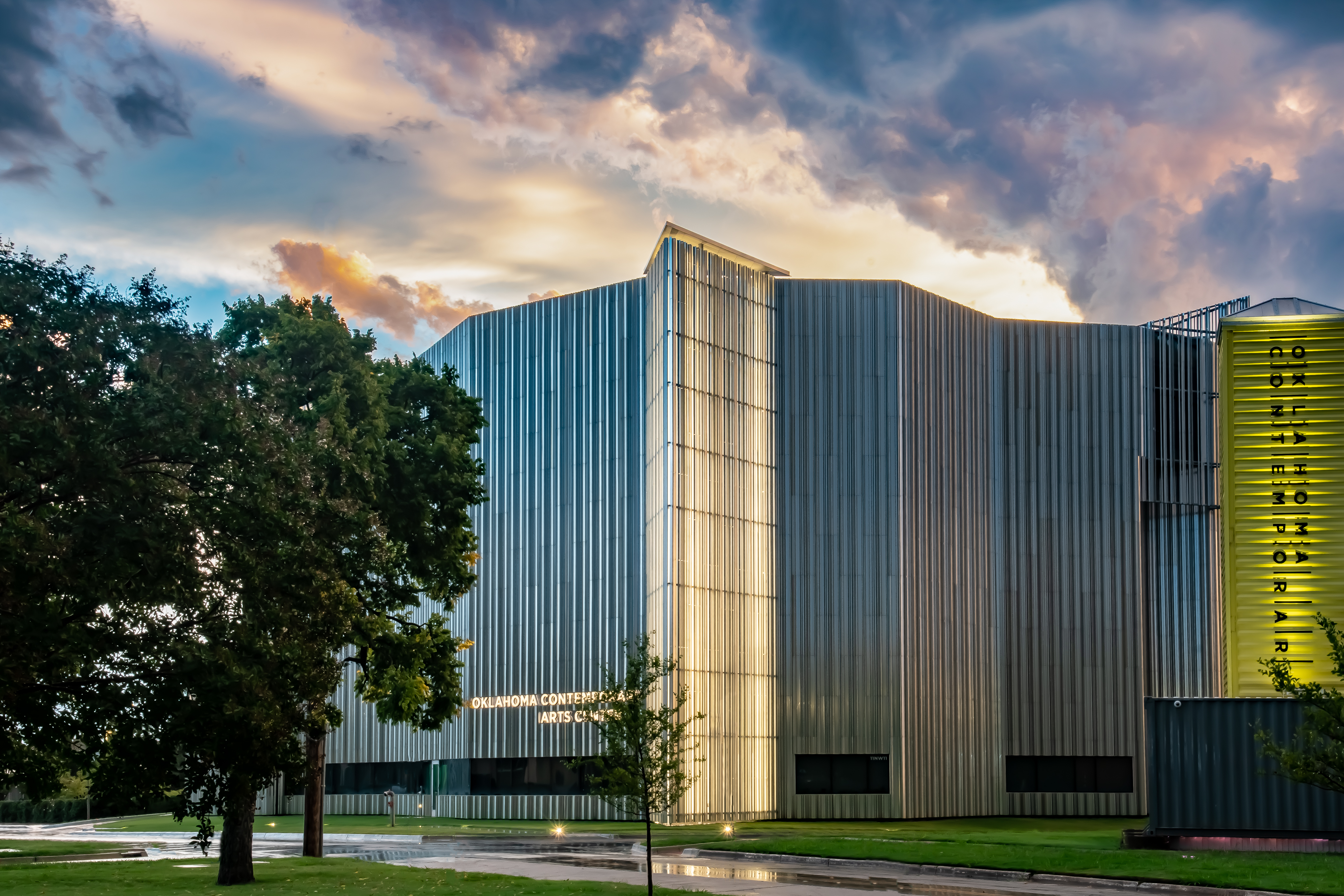 The sky is filled with blue and pink clouds, and the ground looks like there has been recent rain. We can see green grass and full trees in front of a large building covered in aluminum fins reflecting the sky's colors.