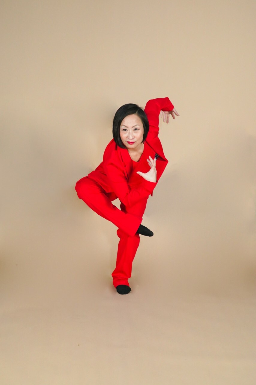 A person in a red suit stands in a neutral background. The person is making a dance pose, folding themselves inward with their arms in different directions and one foot bent over the opposite knee.