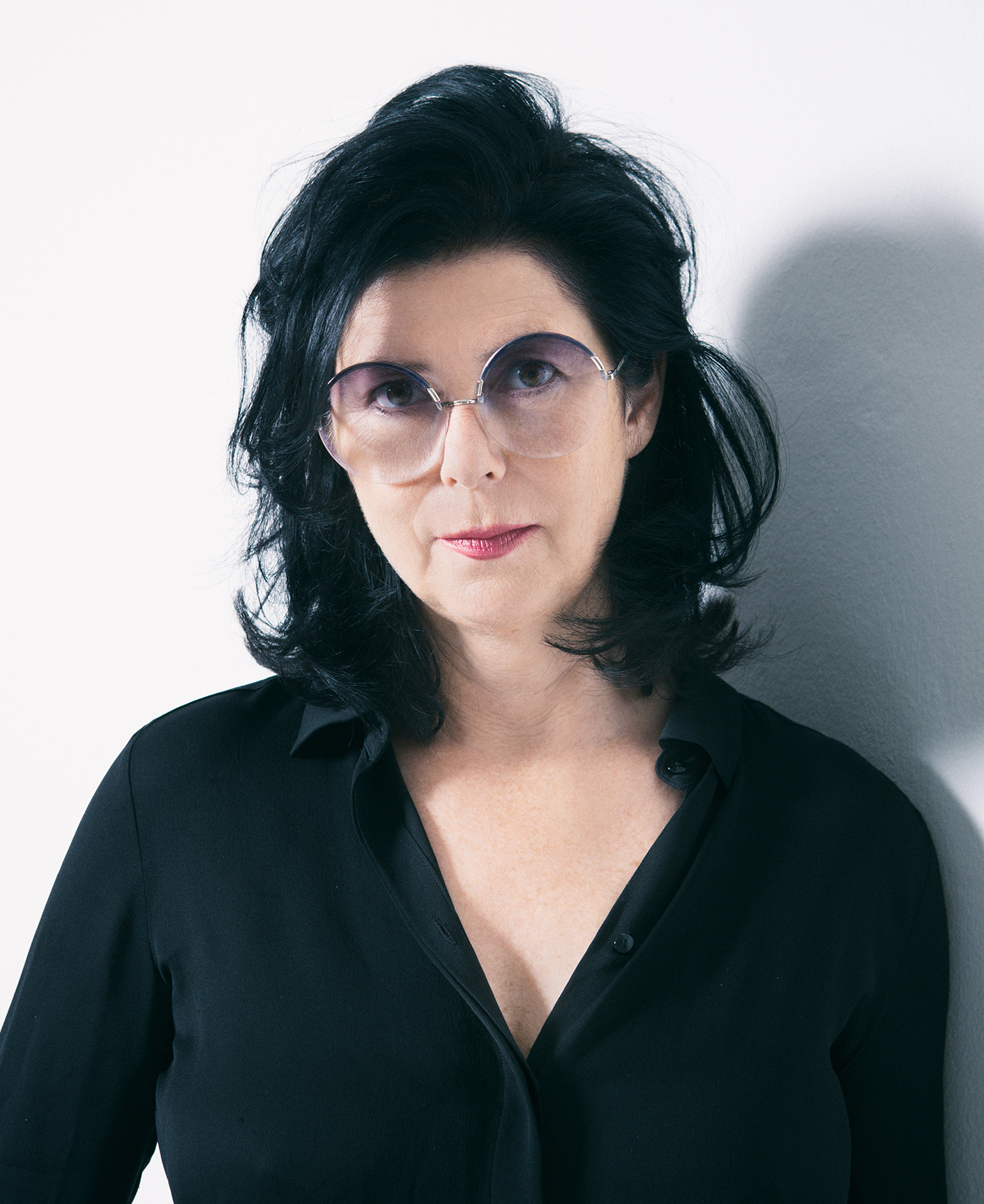 A woman with dark hair and eyes, dressed in a black v-neck, long-sleeve shirt and wearing gray-tinted glasses is looking at the camera.