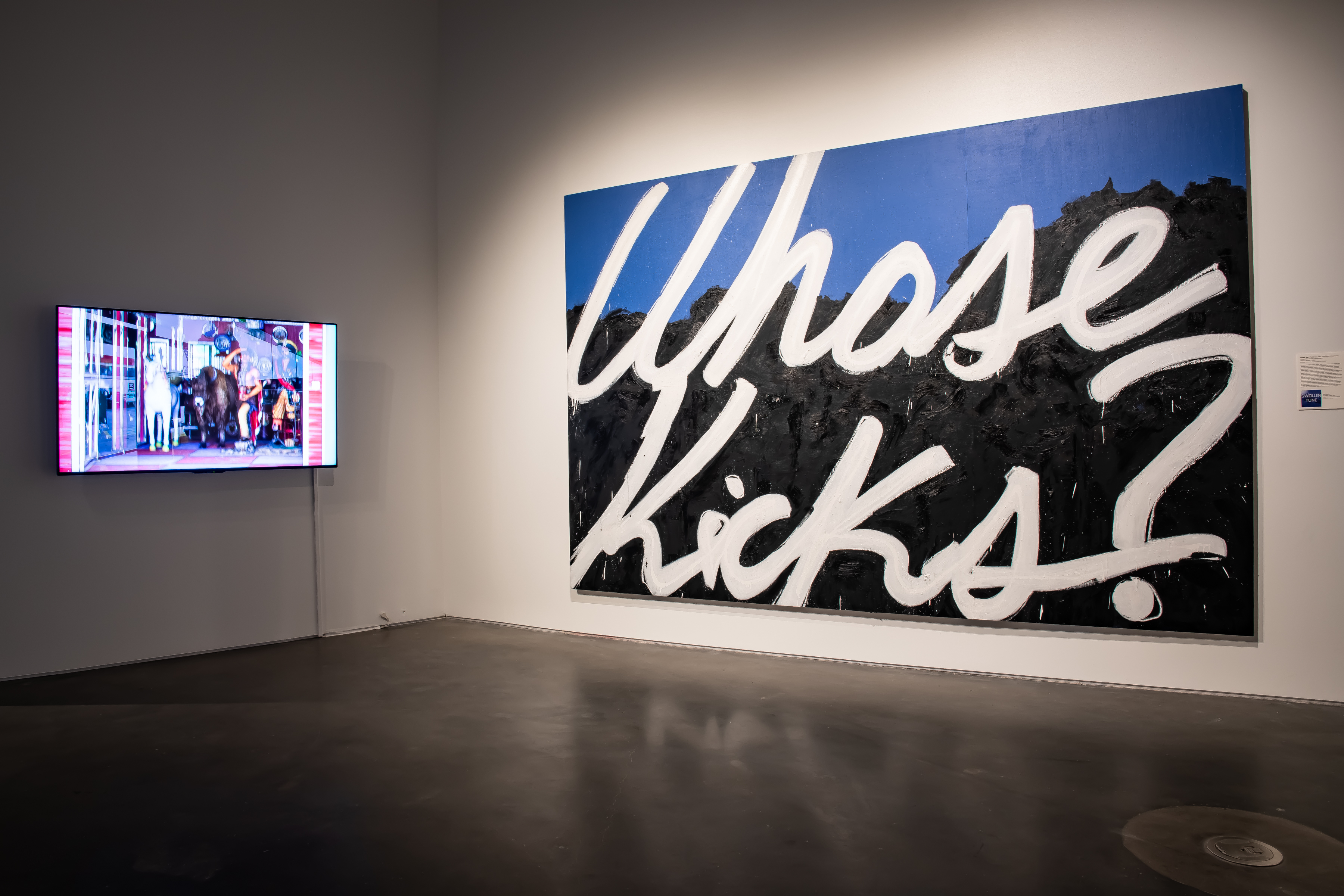 Two works of art hang on a white gallery wall. Left: A video work on a TV. Right: A large blue and black mural with the words WHOSE KICKS? painted in white in the center.