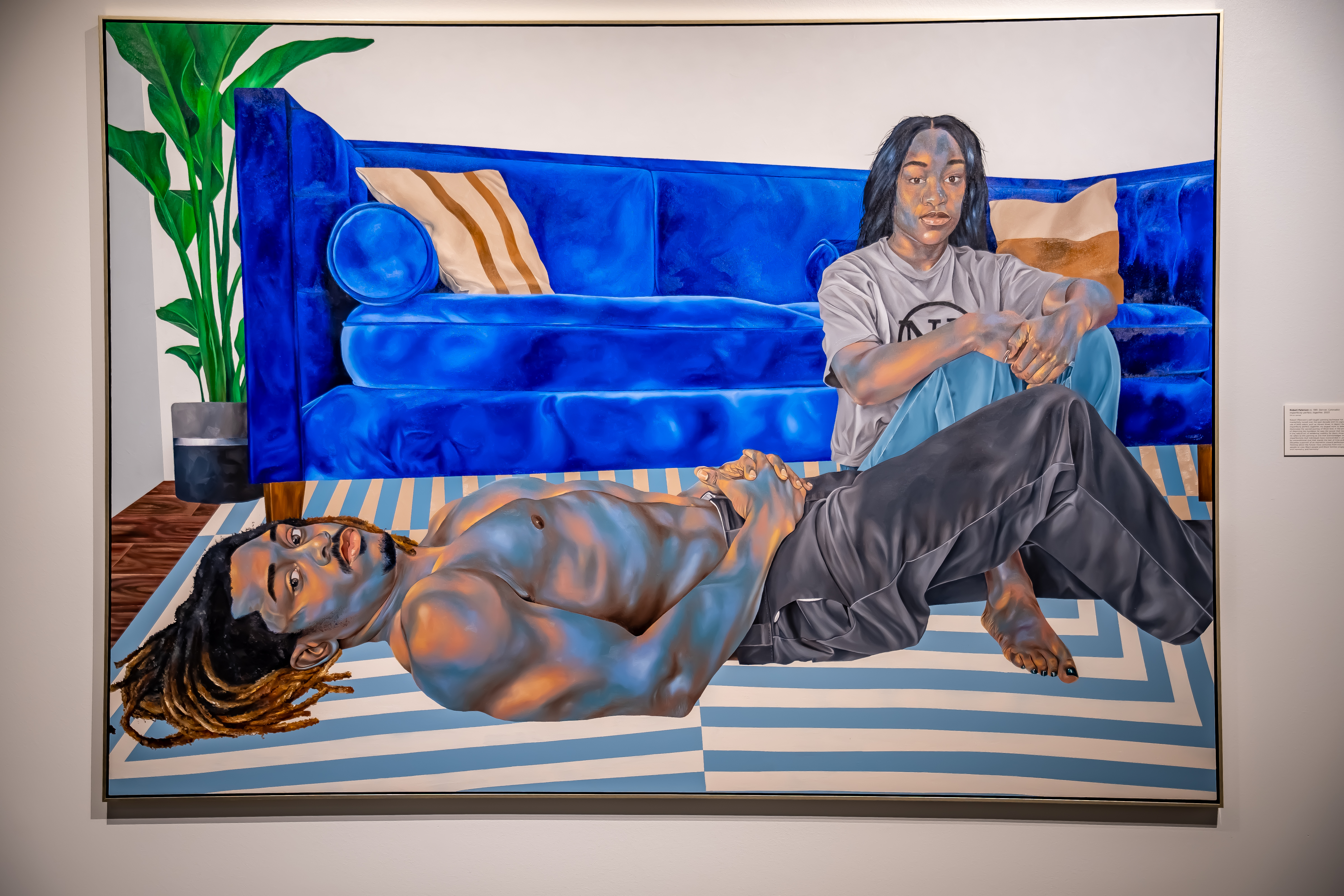 A painting shows two Black people lounging on a blue and white striped rug in front of a stark blue sofa. They are accented in blue and peach tones.