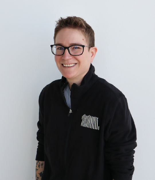 A white person with short hair, dark rimmed glasses and dressed in a black pullover stands in front of a white wall smiling