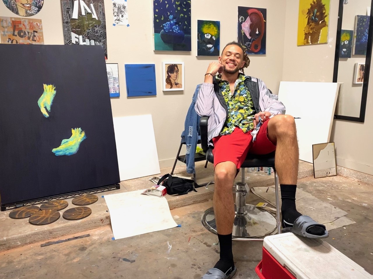 A smiling figure in colorful clothing sits back in a chair in an art studio with paintings on the wall
