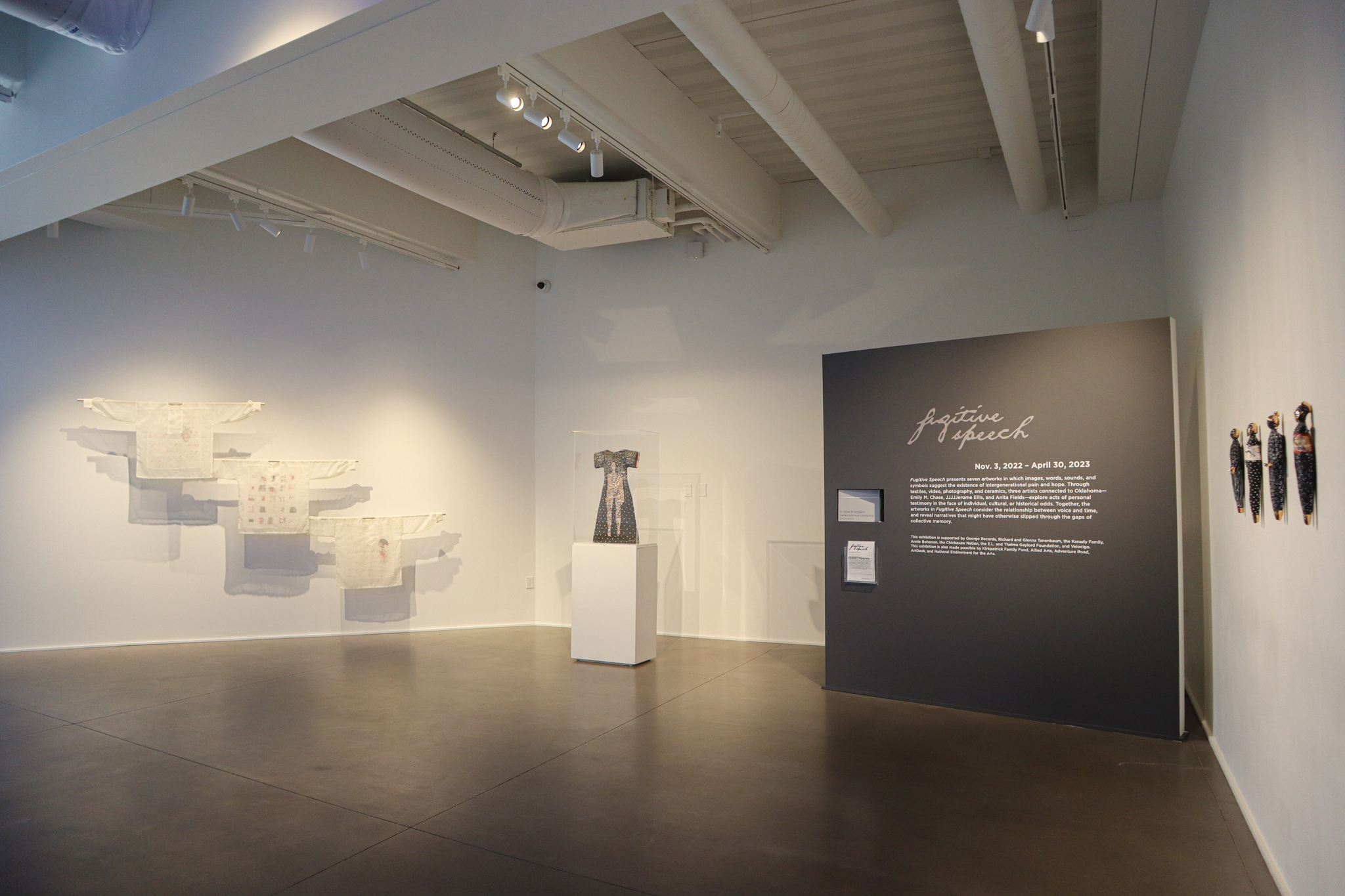 Fiber and ceramic artworks hang and stand in a gallery with a gray wall that says in cursive "Fugitive Speech"