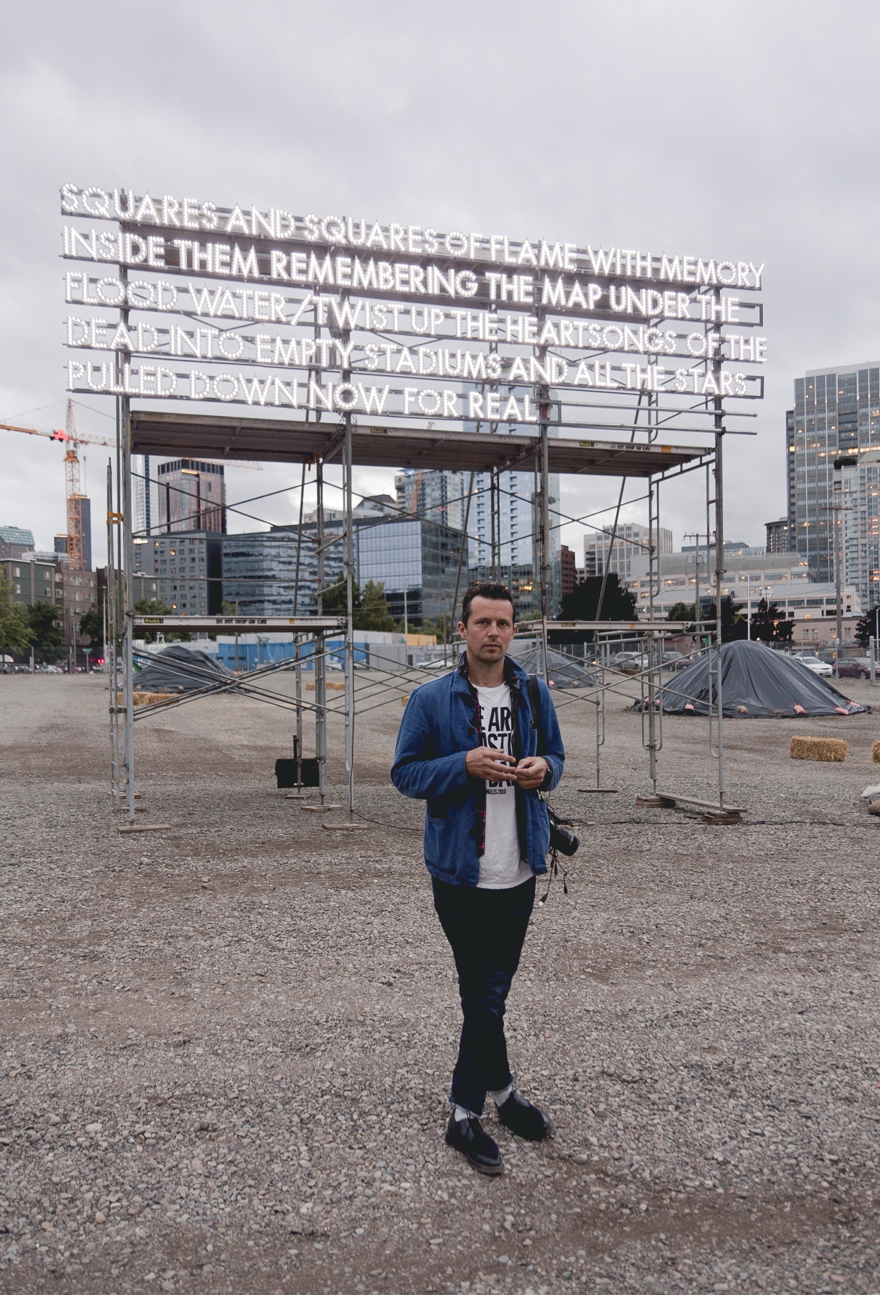 A person stands in front of a work of billboard installation art with a skyline looming in the background