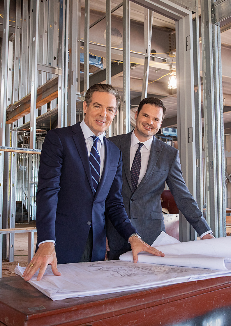 Two men in suits stand in a building under construction and look at plans on a table