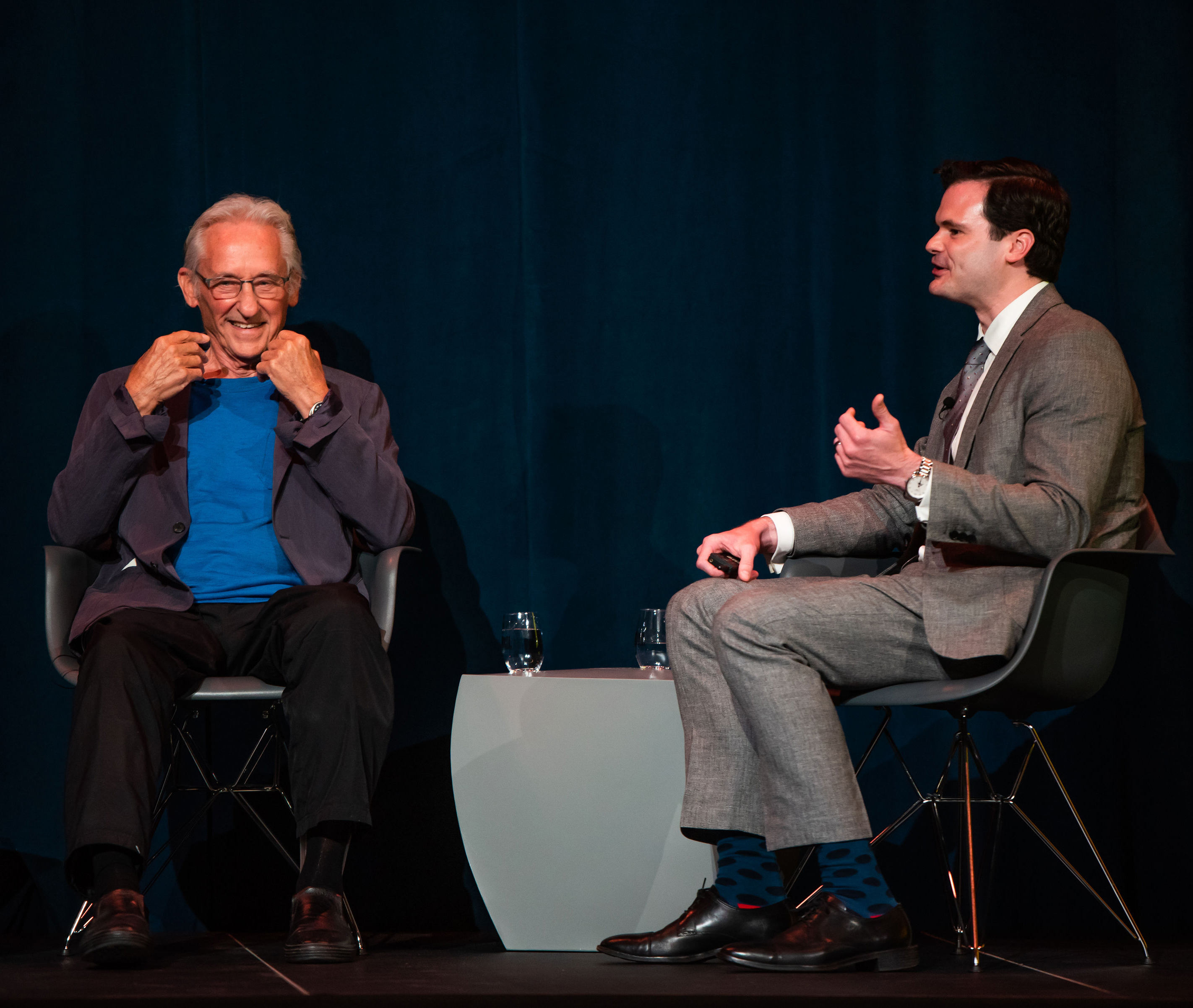Two men -- one in a blue shirt and sport coat and one in a suit -- talk on a stage