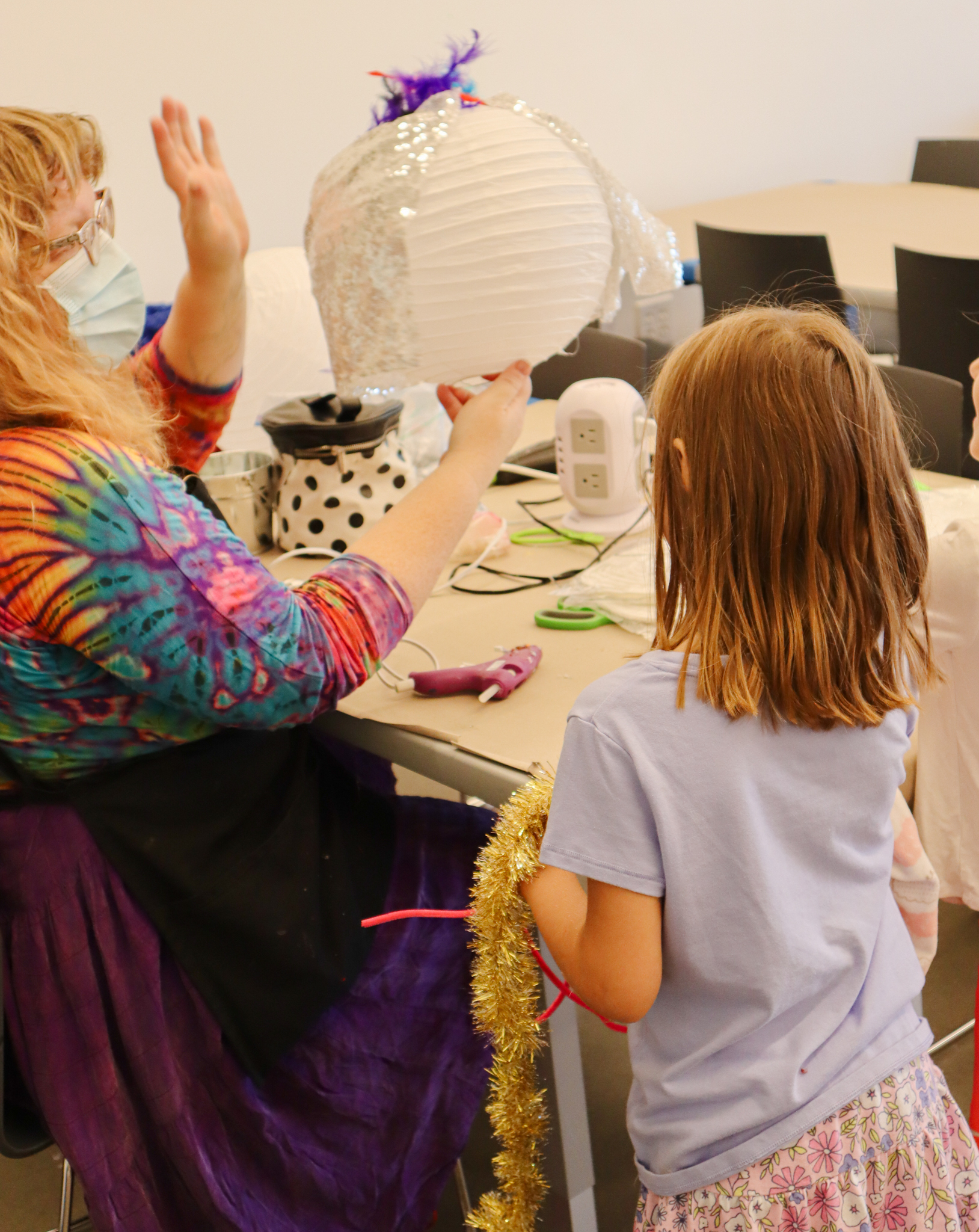 A person in a tie-dyed shirt and purple skirt holds up a circle lantern with purple feathers and a sparkly cape. A little kid in a purple shirt and floral skirt stands in front.