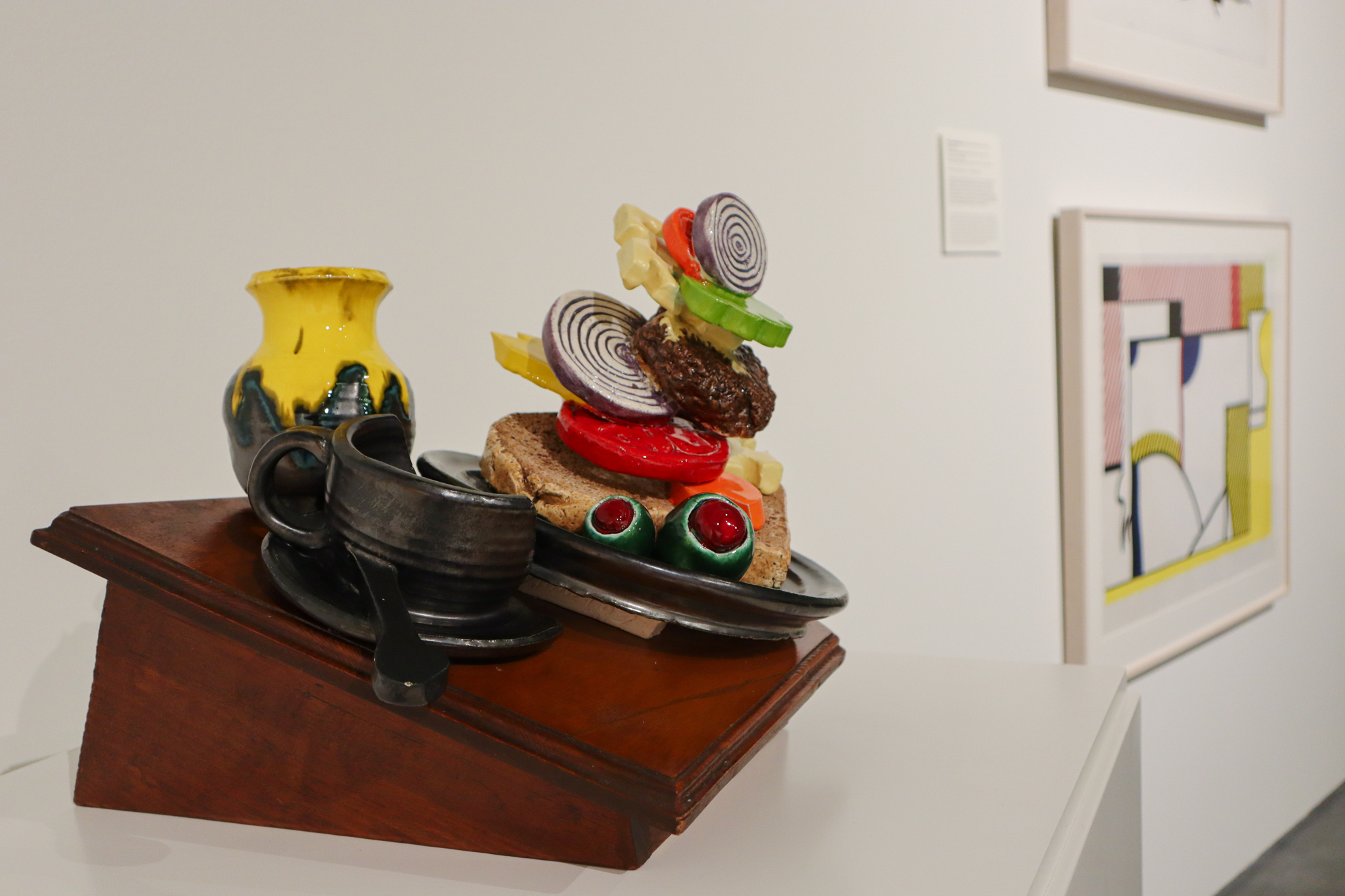 A ceramic sculpture is in frame. In Pop Art style with bright colors, we see a mug with coffee in it, a jar with what looks like mustard coming out of the top, and a deconstructed sandwich with a patty, onion, lettuce, cheese, tomato and olives on a plate