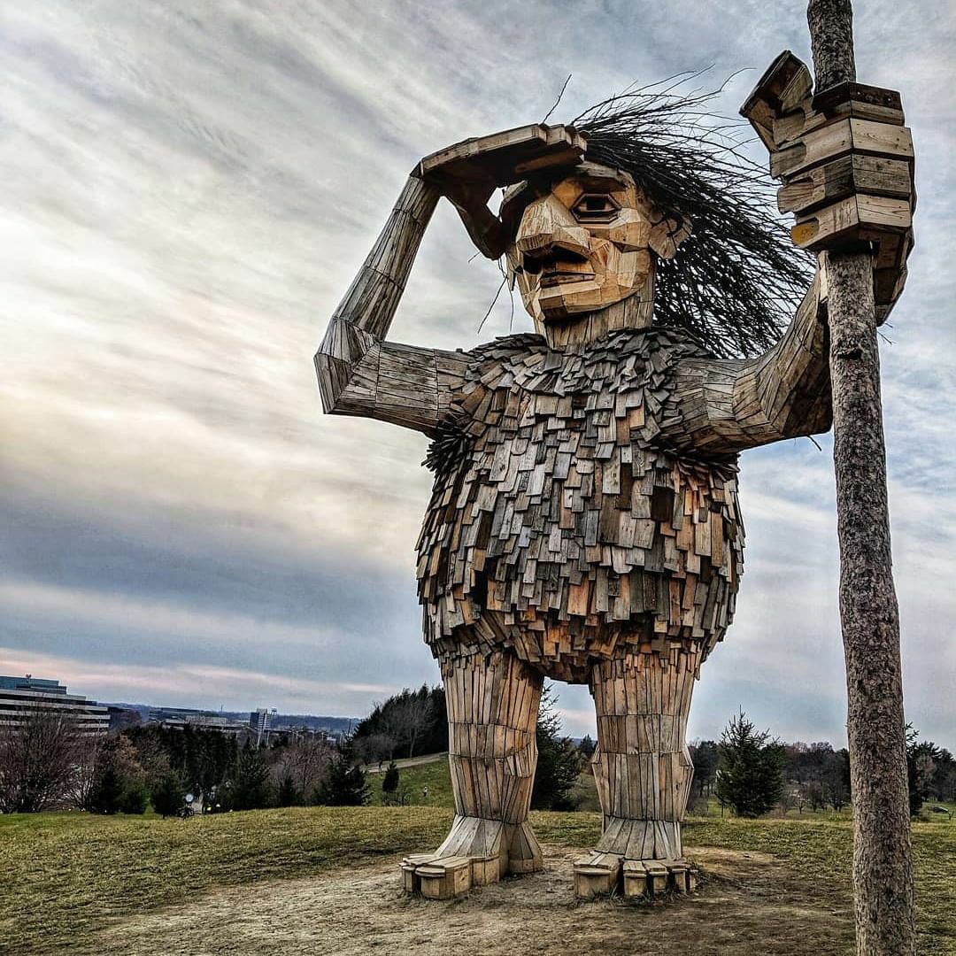 A large, wooden, troll-like sculpture stands on two feet, with a thick torso, long arms and large hands, a big nose and wild straw-like hair. The sculpture is outdoors, and one hand is over its right eye as it holds a staff in the other hand, looking out.
