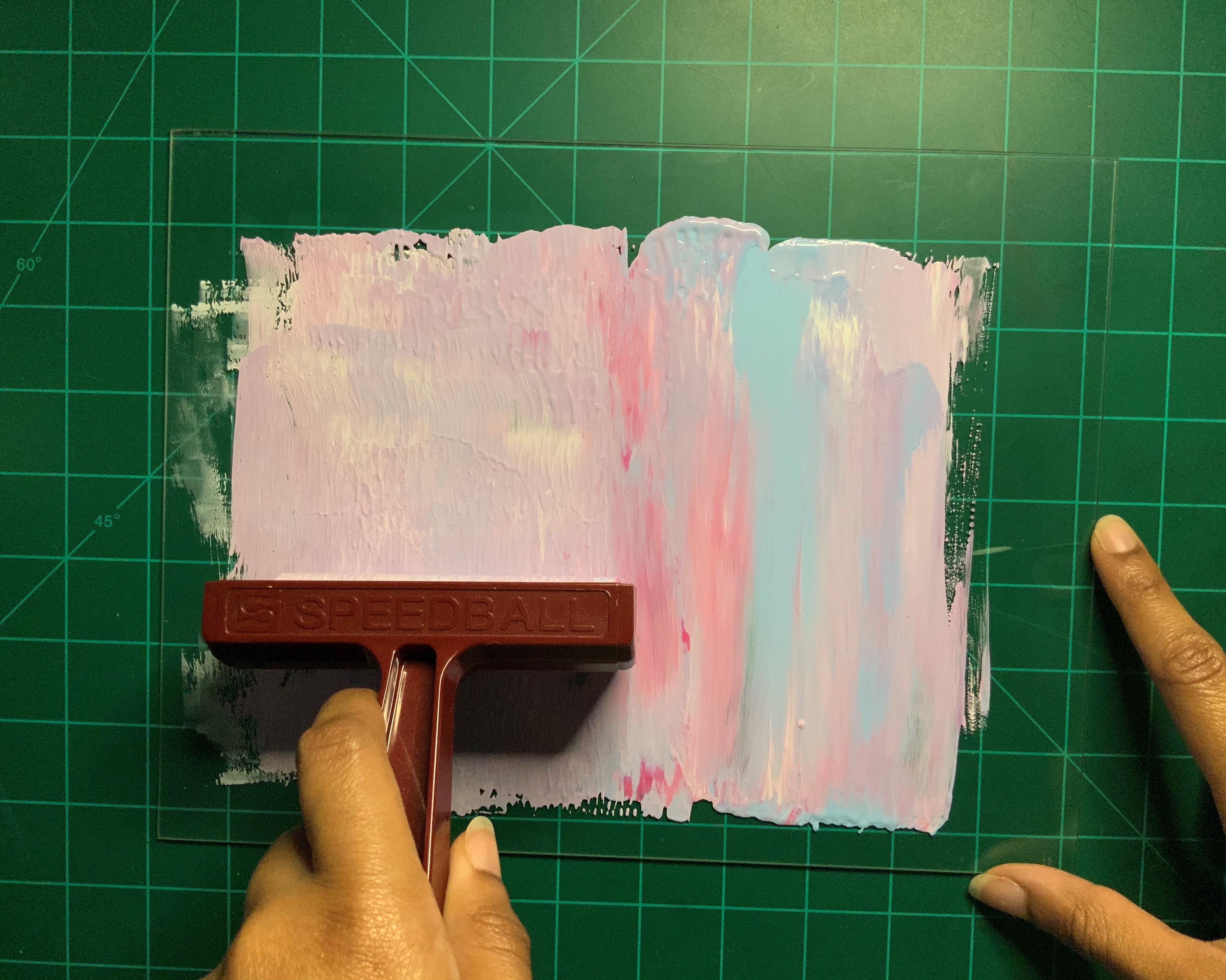 A hand rolls out pastel pink and blue paint using a brayer