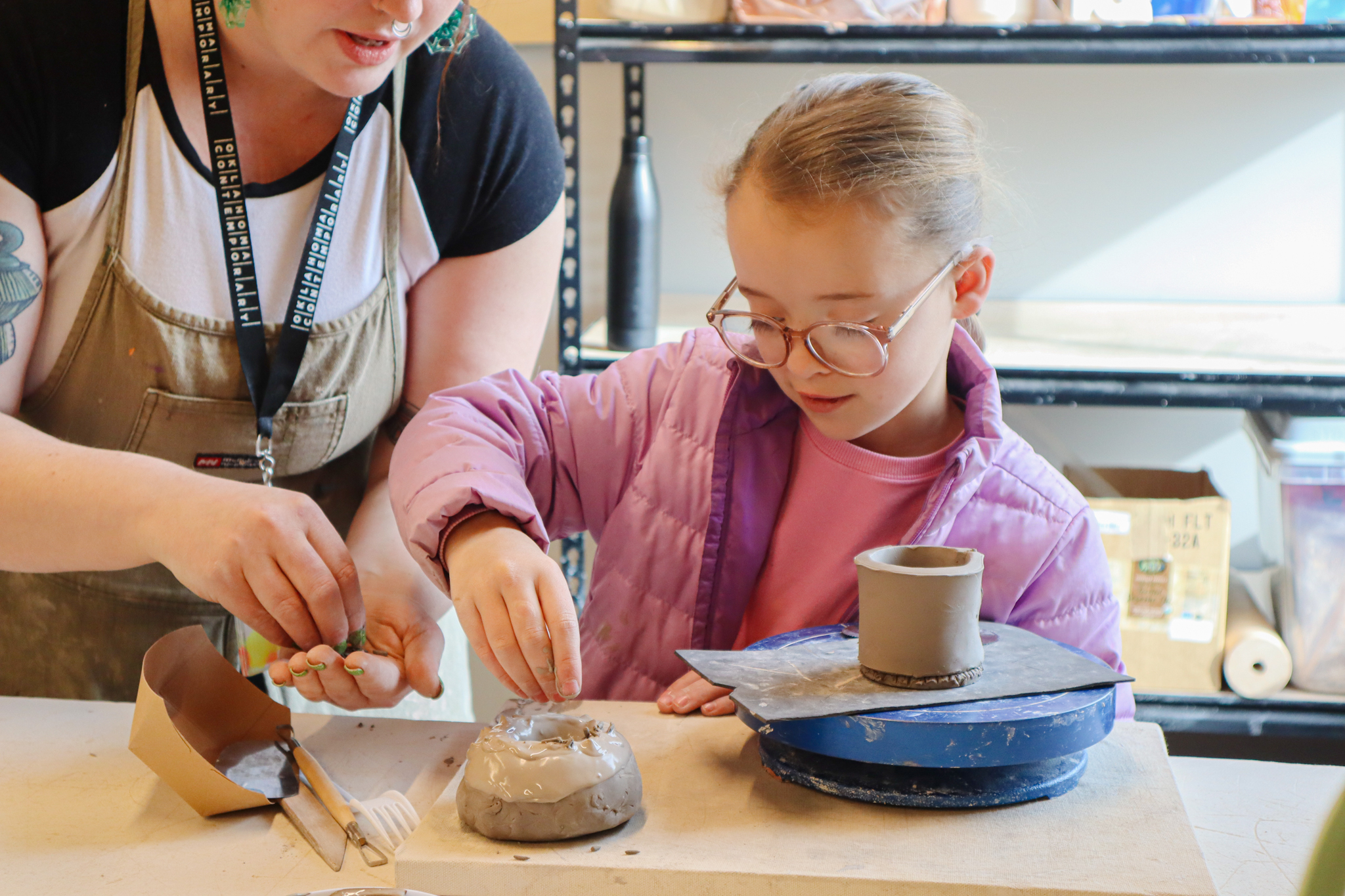 A kid is sprinkling clay sprinkles onto a clay donut. They have round glasses, a pink shirt and purple jacket on. Someone is leaning in to help them.