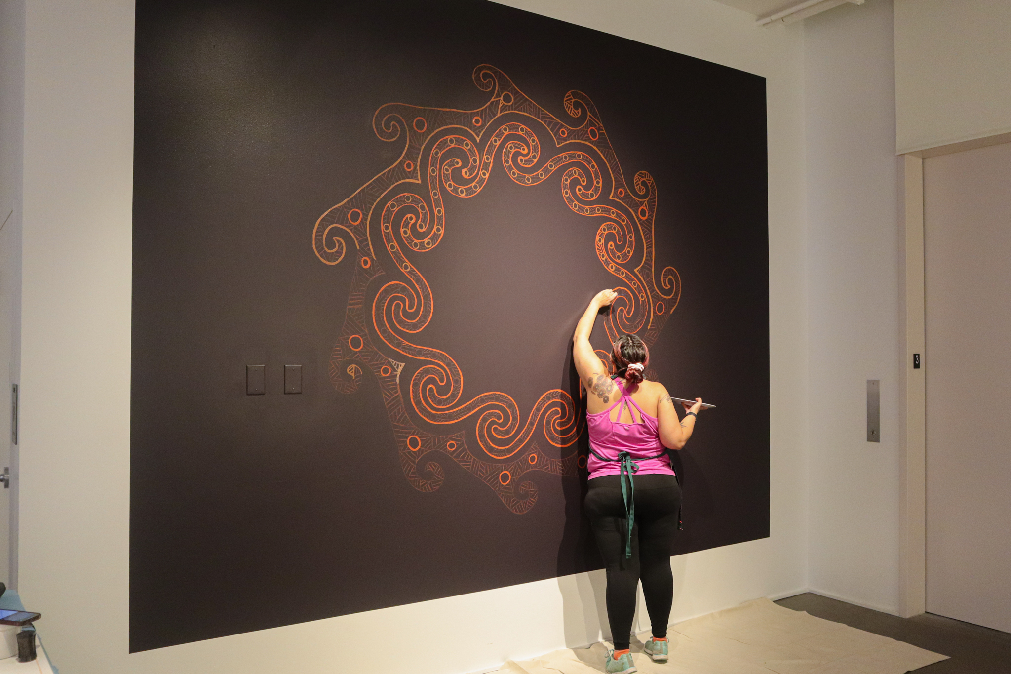 A person is painting on a large black sqaure painted on a white wall. The person has a pink top and black leggings on with dark hair pulled back. They are painting a swirling star-like pattern in deep reds and oranges.