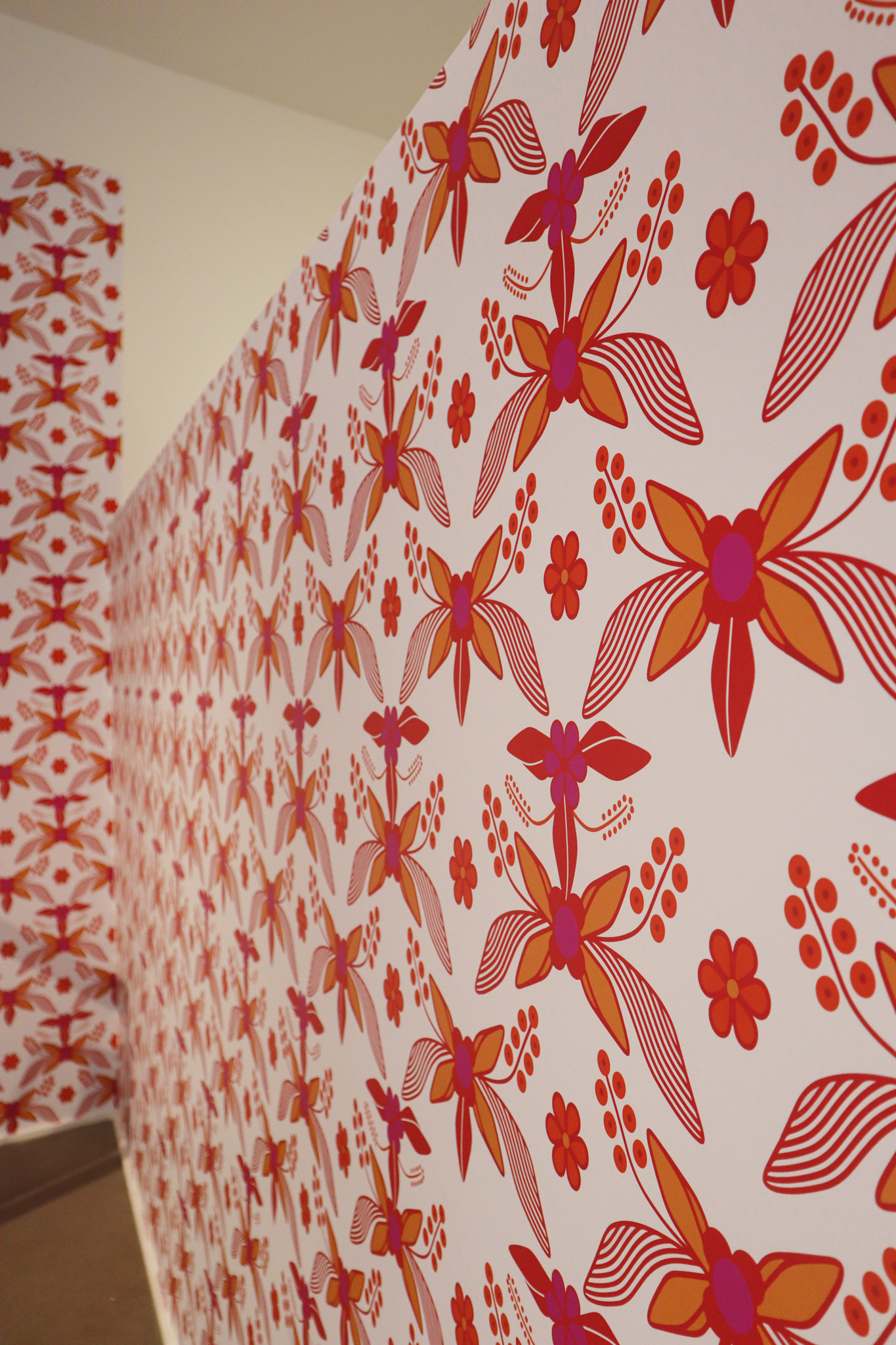 Colorful wallpaper sprawls across a white wall. Looking at an angle, we can see bright pink, red and orange patterns of what looks like a butterfly/flower creature, replicated over and over again.