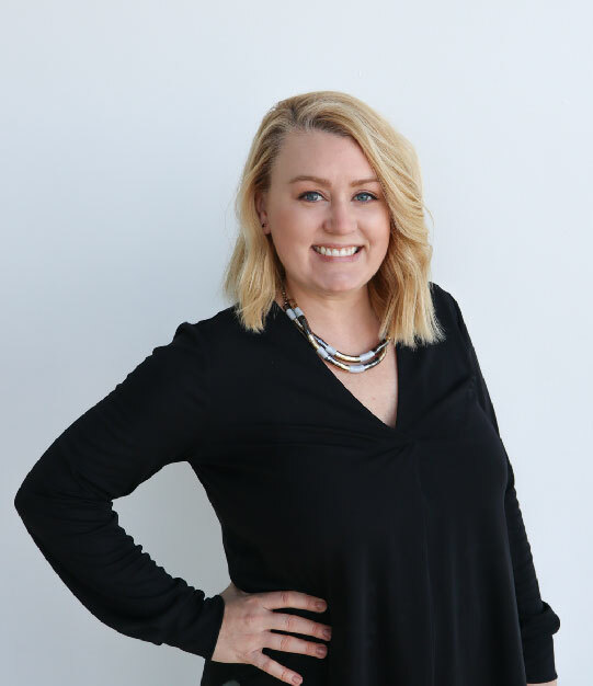 A white woman with short blonde hair, a thick necklace and black long-sleeve shirt stands with her right hand on her hip in front of a white wall, smiling