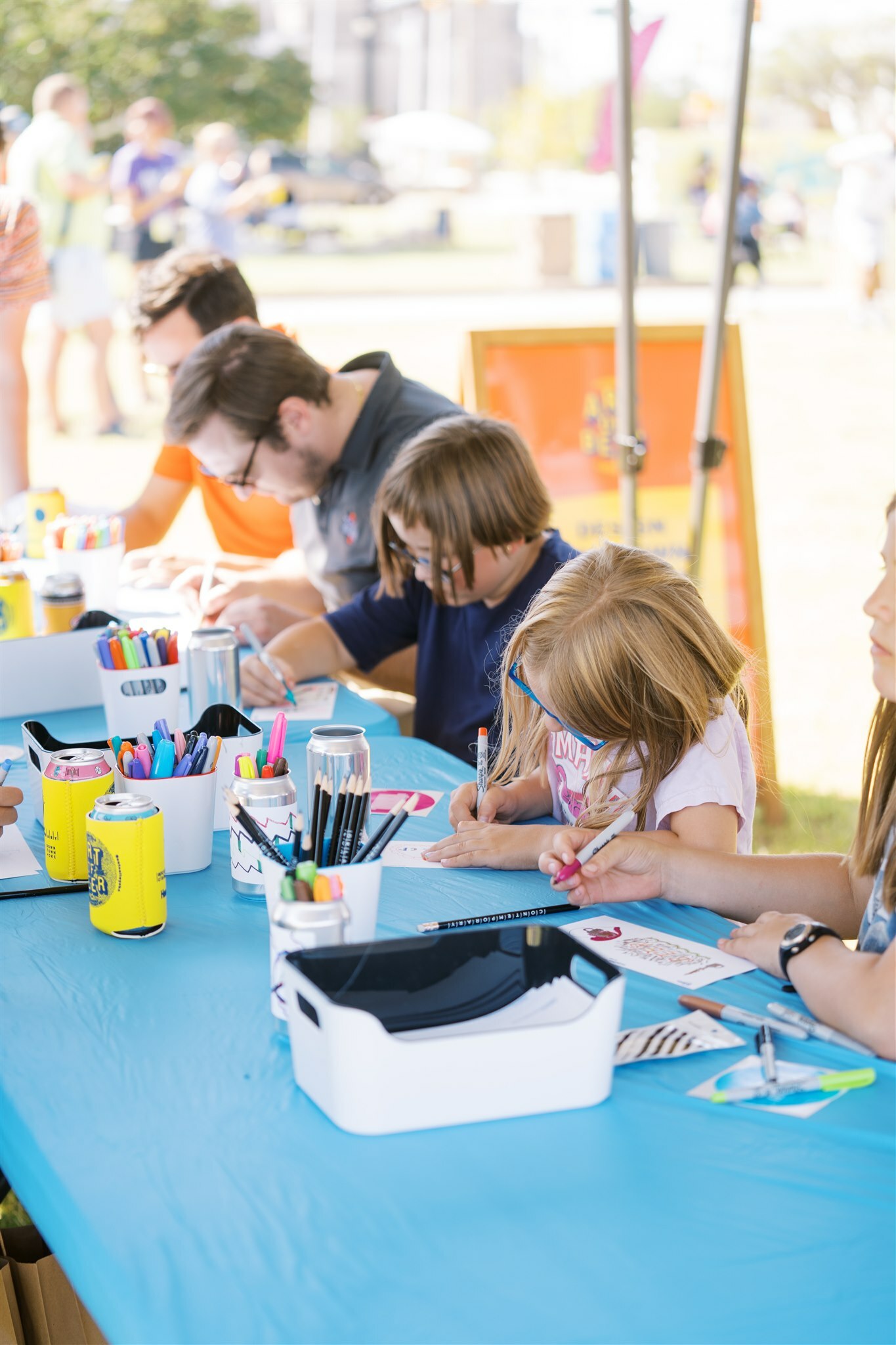Kids and adults alike are sitting a table with a blue tablecloth. There are boxes and cups of markers and pencils on the table. Everyone is art-making.