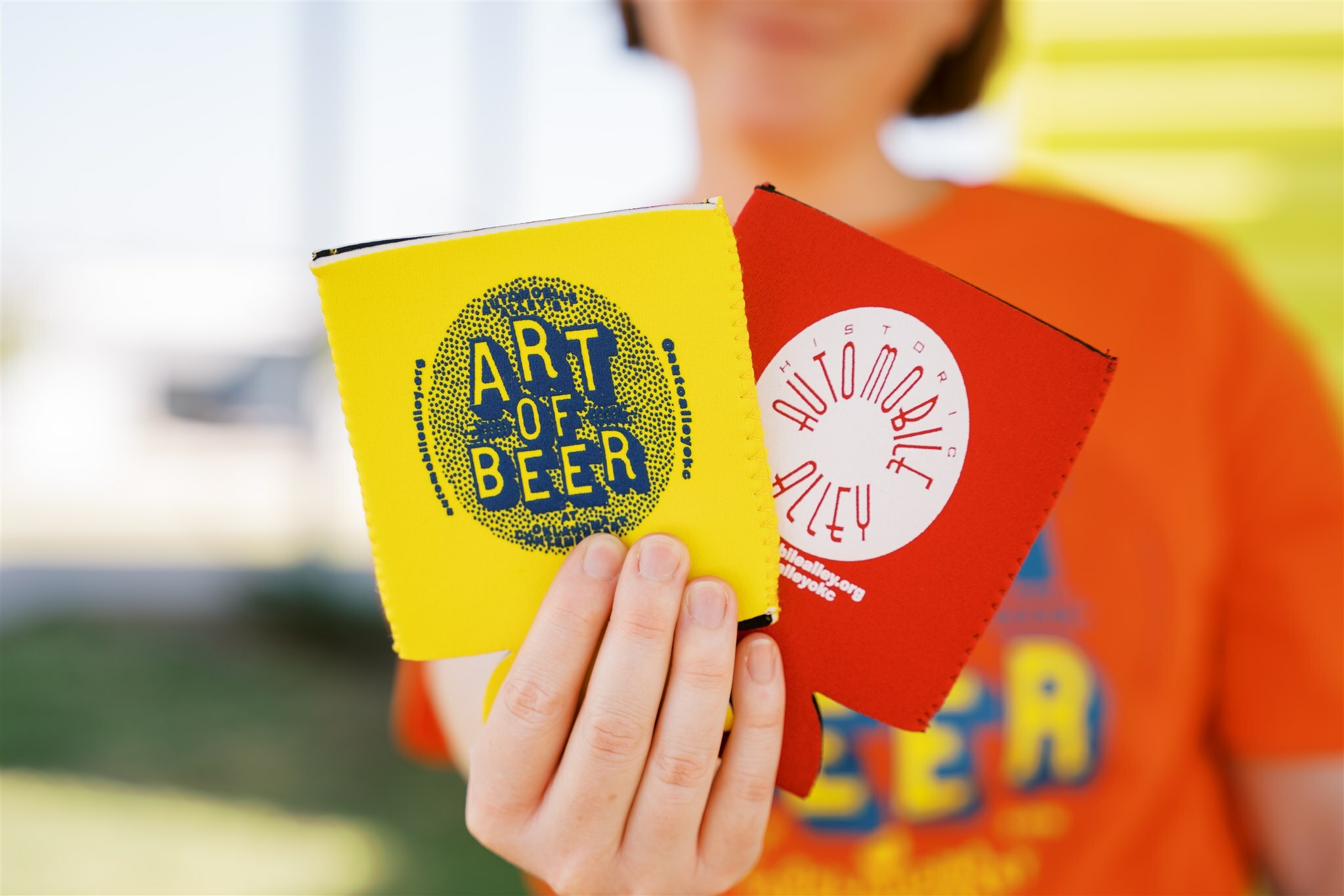 A person is holding two koozies. One is yellow and has 'Art of Beer' printed in a blue circle. The other is red and has 'Automobile Alley' printed in red in a white circle, forming a circle with the words.