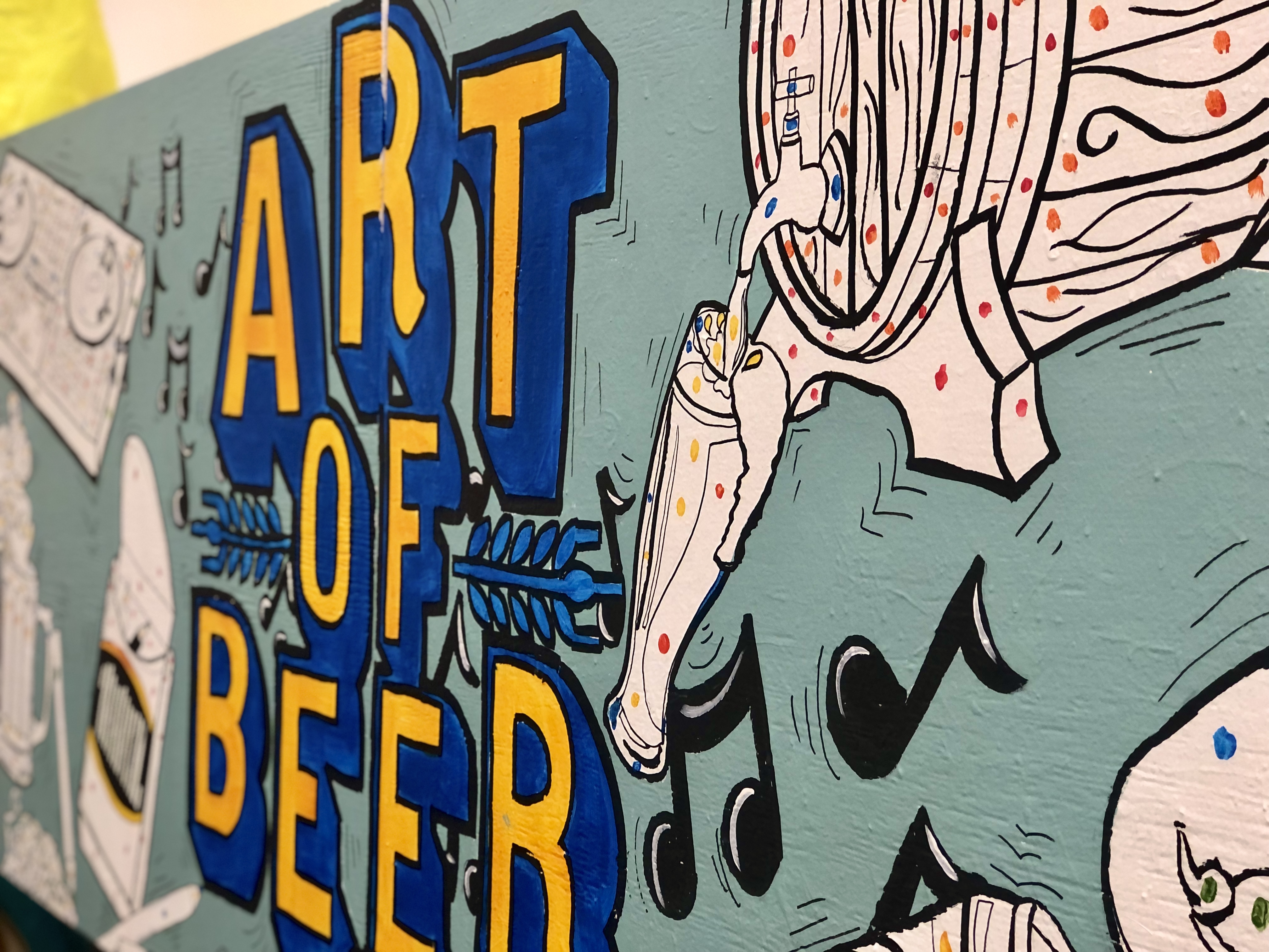 A large mural is painted on a mobile wall. The abckground is a light blue, with Art of Beer painted in the middle in yellow and dark blue. Surrounding the logo is outlines of beer glasses, a turntable, wheat and barley and musical notes.