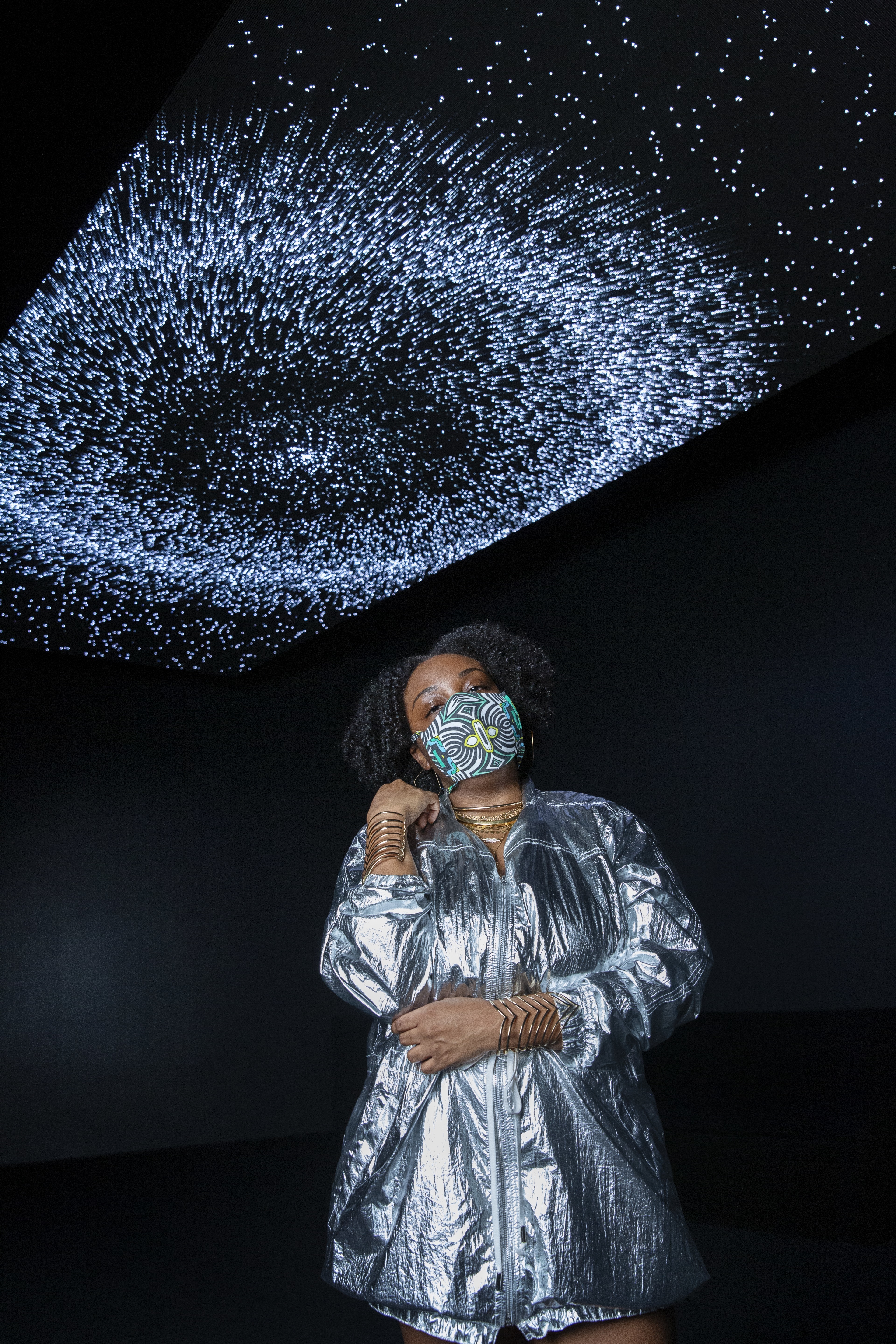 A masked figure poses in front of a digital art installation mimicking a starry night sky