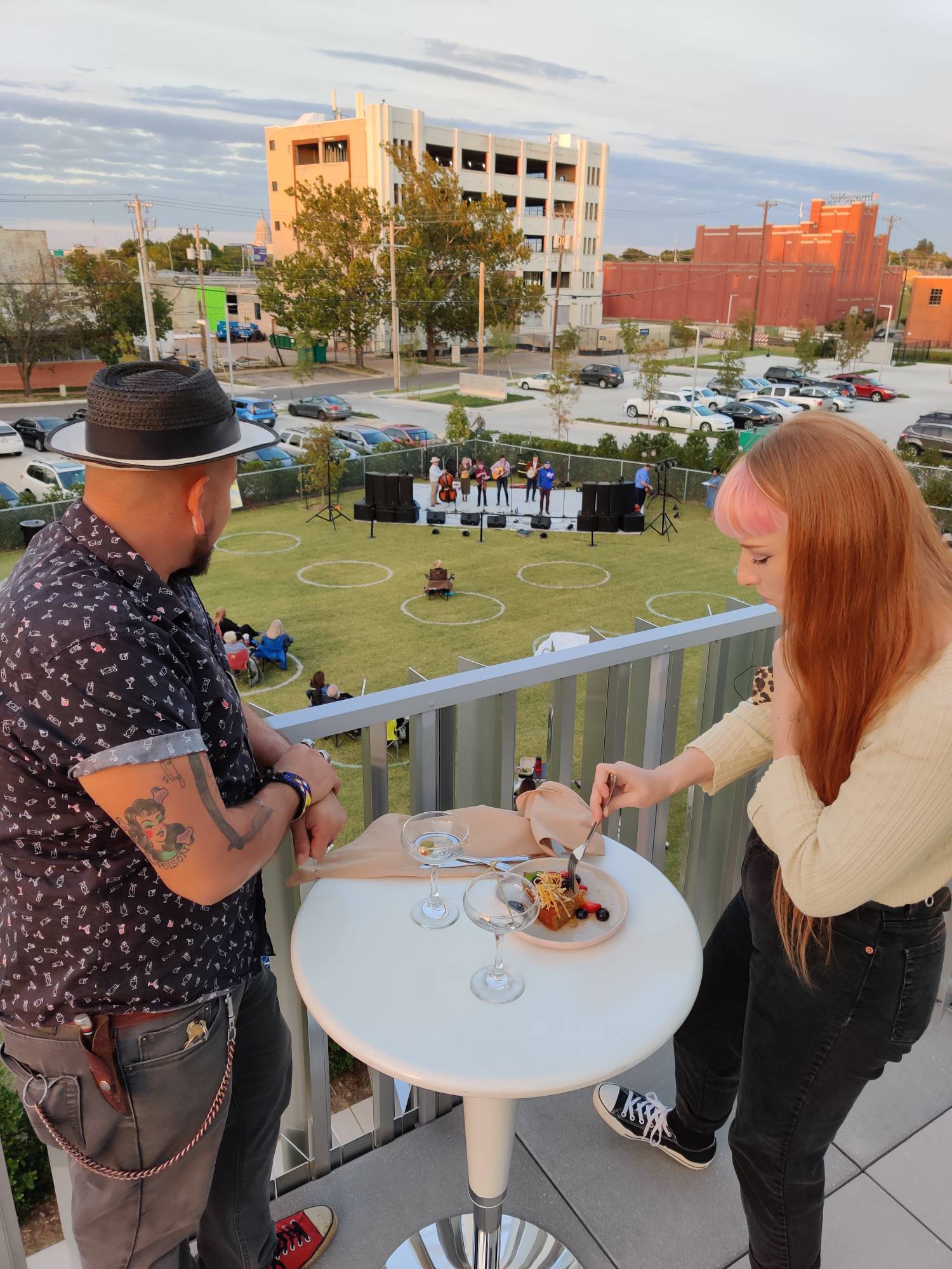 Two people eat outdoors on a patio overlooking a concert on a green lawn