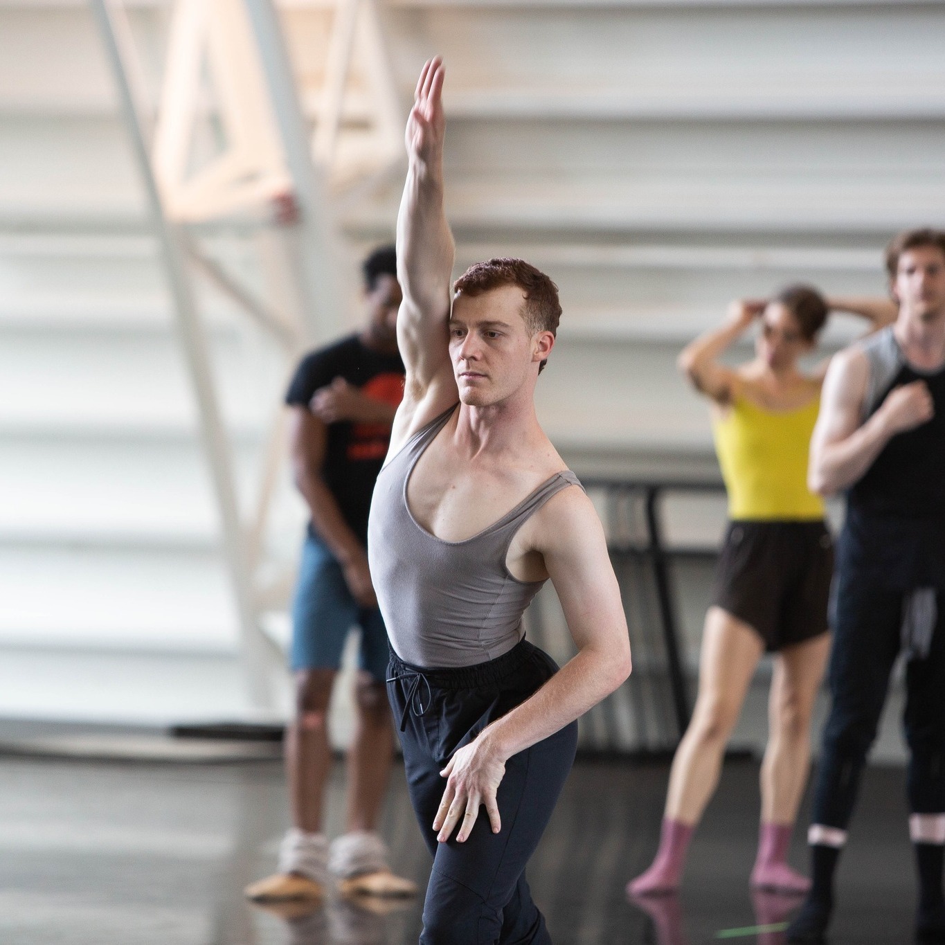 A ballet dancer in a grey top extends an arm overhead. We can see other dancers surrounded in the background.