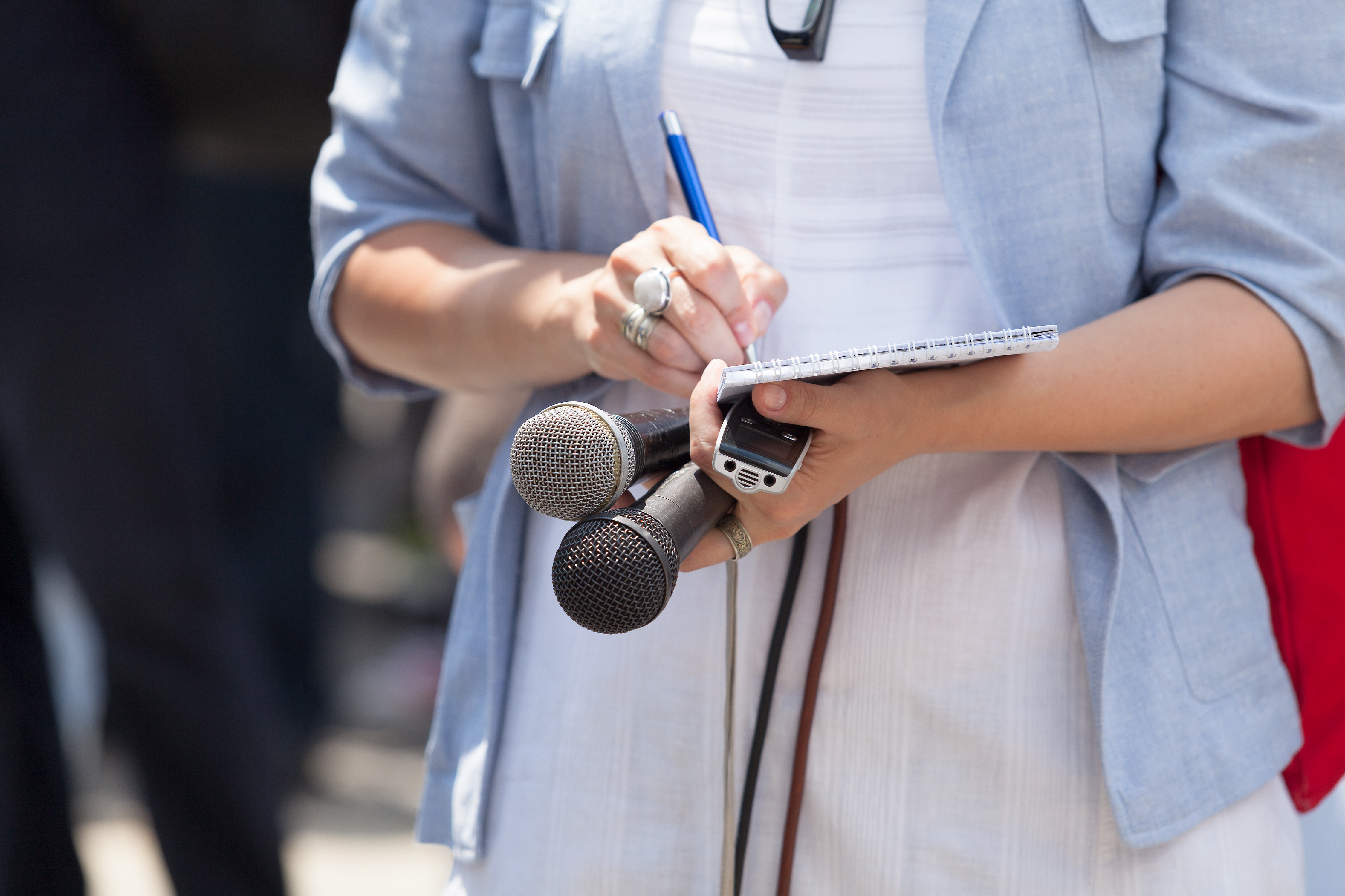 A cropped photo features the torso of a person holding microphones and writing in a spiral notebook