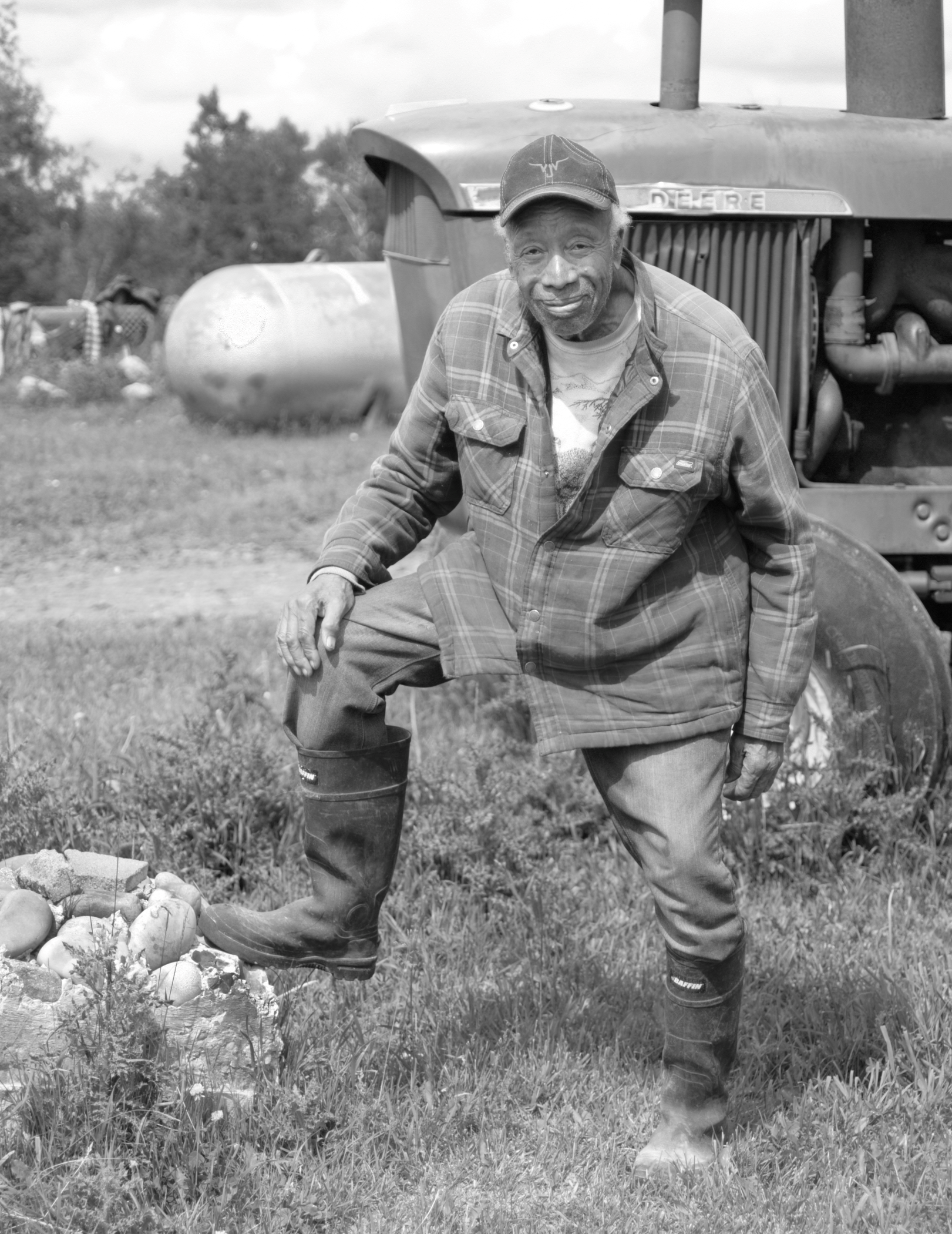 A Black person wearing rubber boots and a ball cap stands with one foot on a rock. A tractor and gas tank are visible in the background.