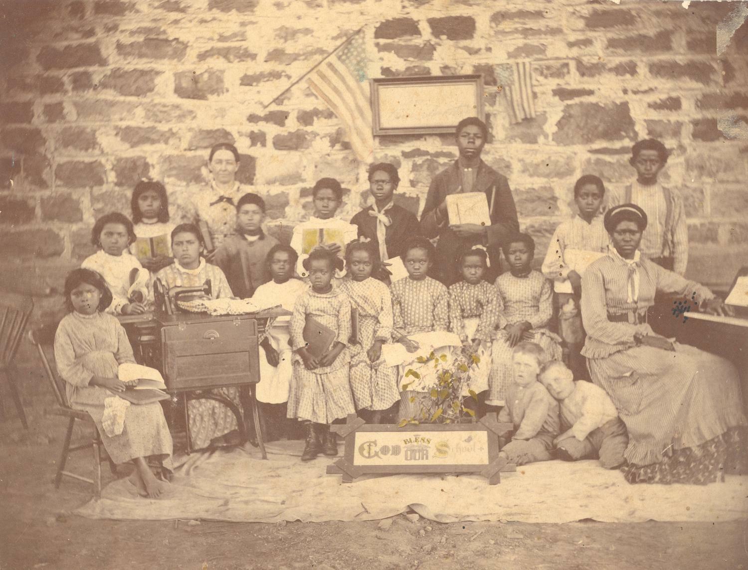 A weathered black-and-white photograph of a group of mostly dark-skinned school children sitting and standing against a brick wall.