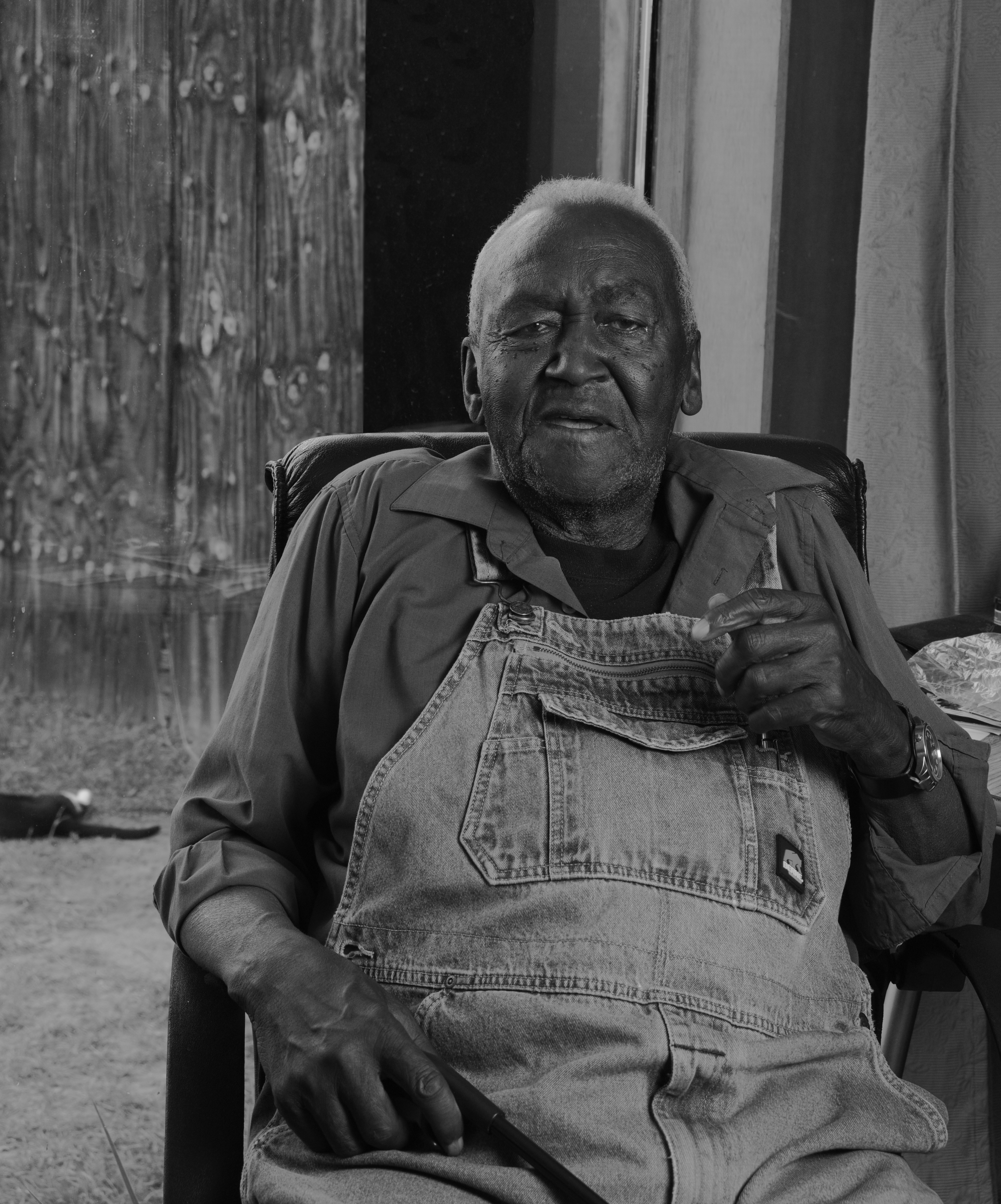 A black-and-white portrait of a Black man sitting down, from the waist up. He wears overalls and a collard, quarter-length sleeved shirt. His hair is short, and his left hand is raised as he speaks.