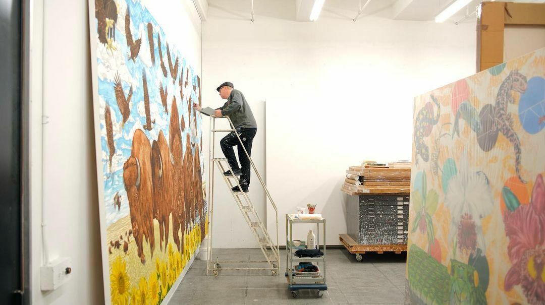A man in paint-covered clothes stands on a ladder at the far edge of a large canvas with charging bisons, swarming eagles and yellow sunflowers painted. The man is leaning forward with a brush in his hand, painting. Behind him is a large painting.