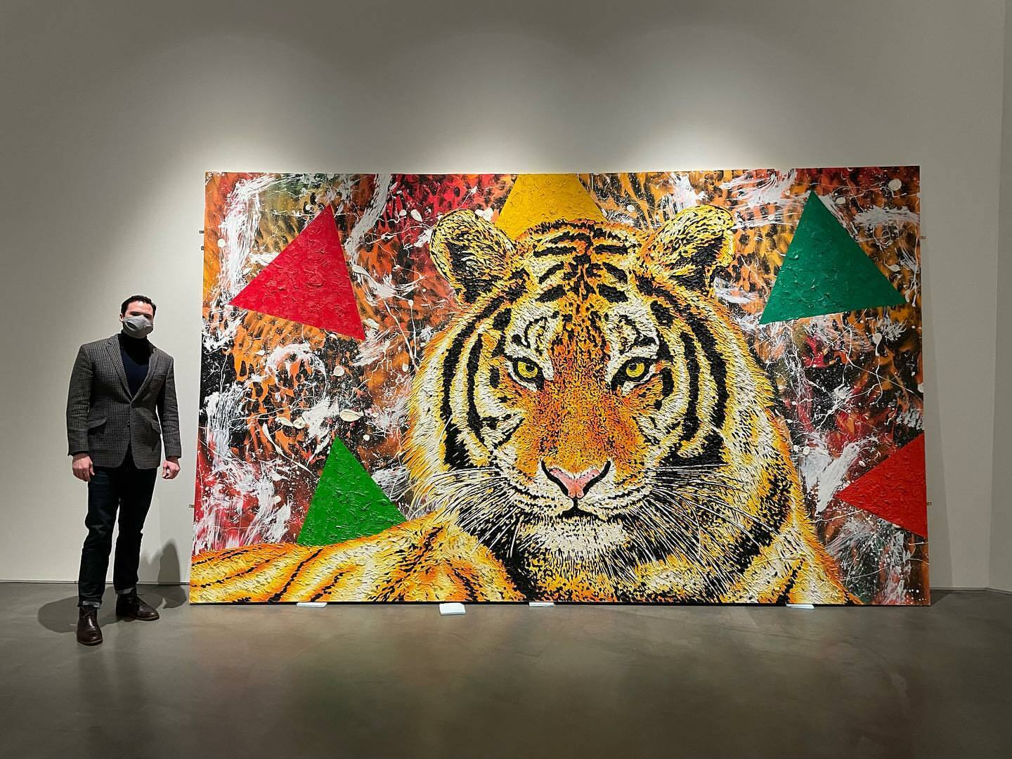 A man in a dark gray suit and black mask stands next to a large oil-painted mural. In the center, a large head of a tiger stares forward with bright yellow eyes. Abstract lines and blotches of paint are in the background with large green and red triangles