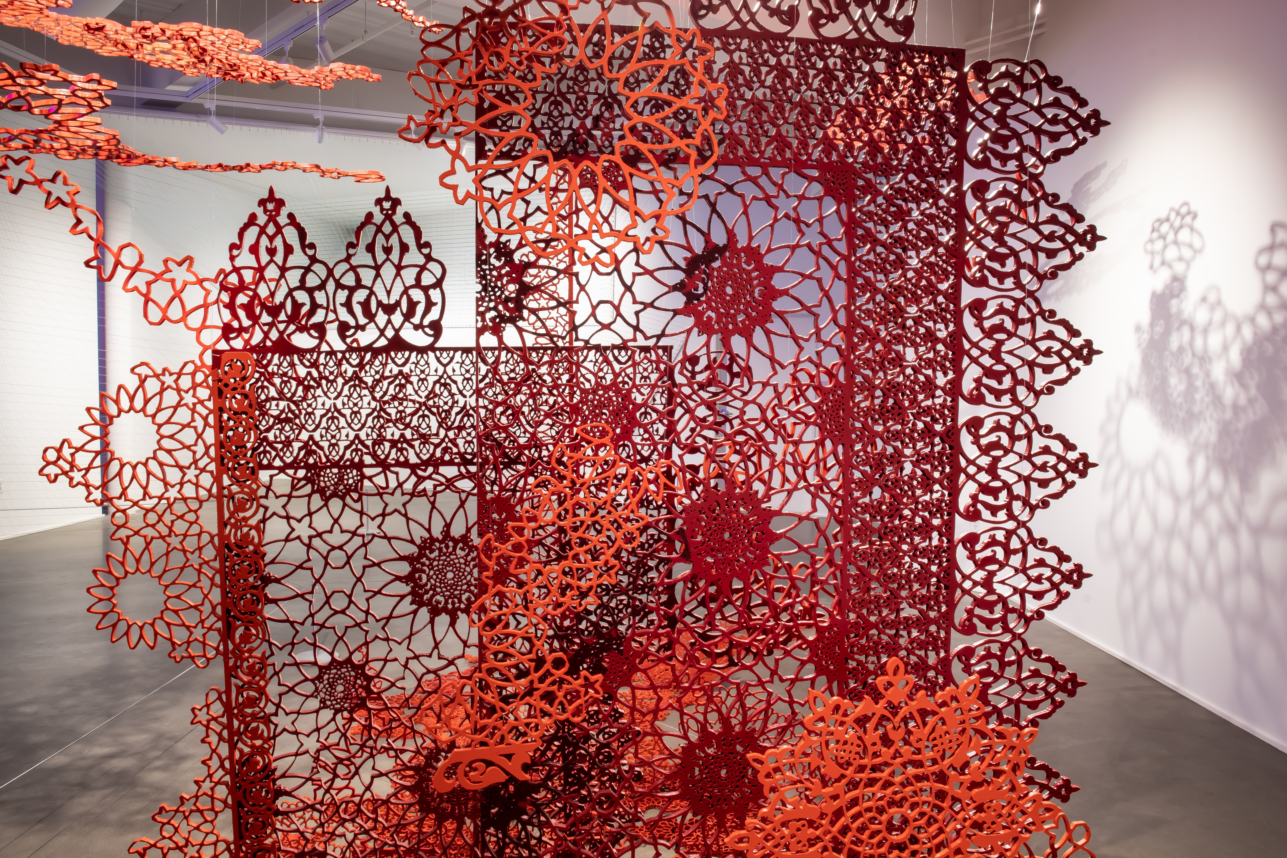 Red, patterned, screen printed pieces stand and suspend from the ceiling, some cut into fragments and some overlapping to create different patterns. There are shadows on the wall, showing similar pattern details from the pieces.