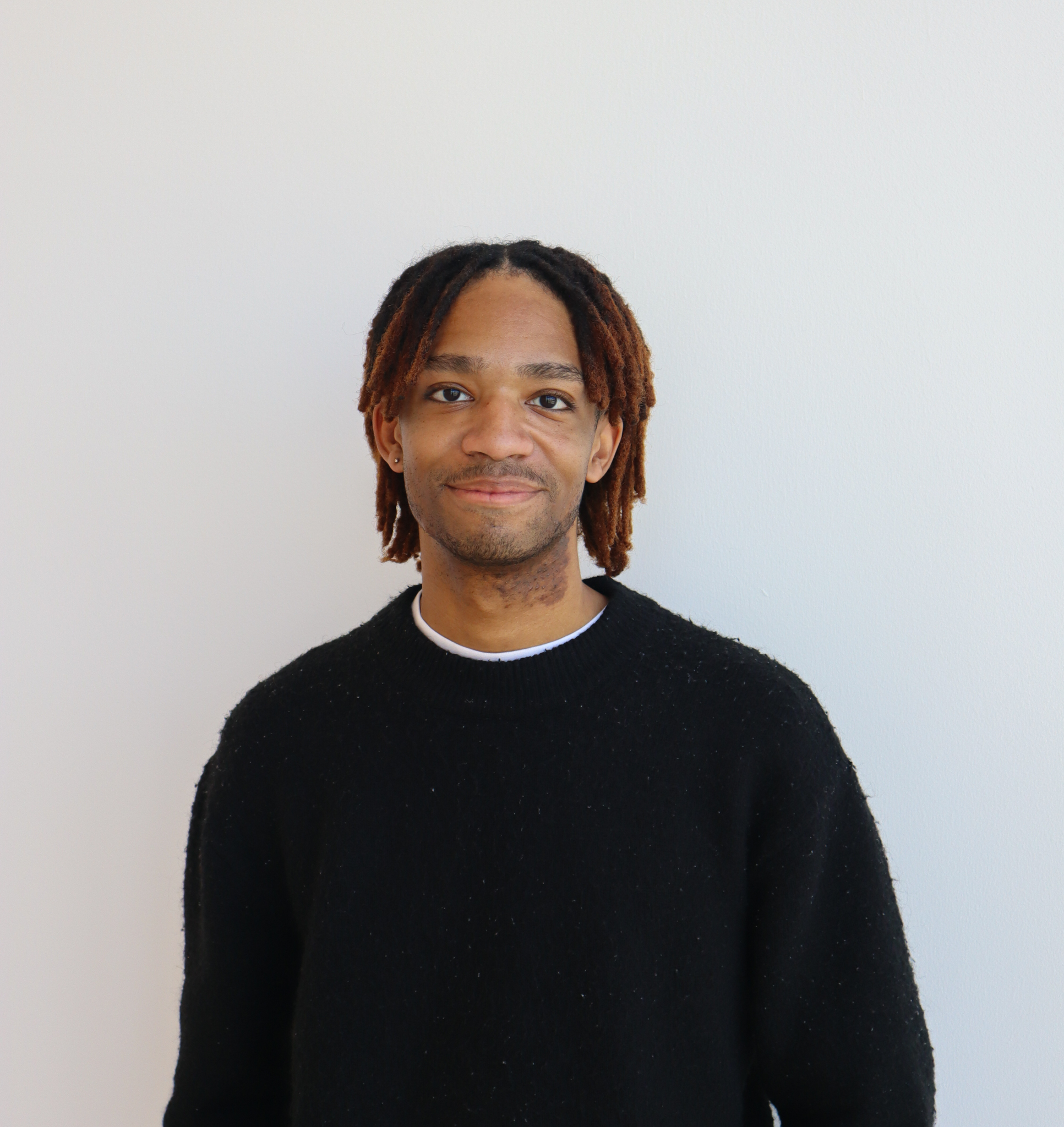 A Black man with short, layered red-brown locs wears a back sweater. He is standing in front of a white wall, smiling softly at camera.