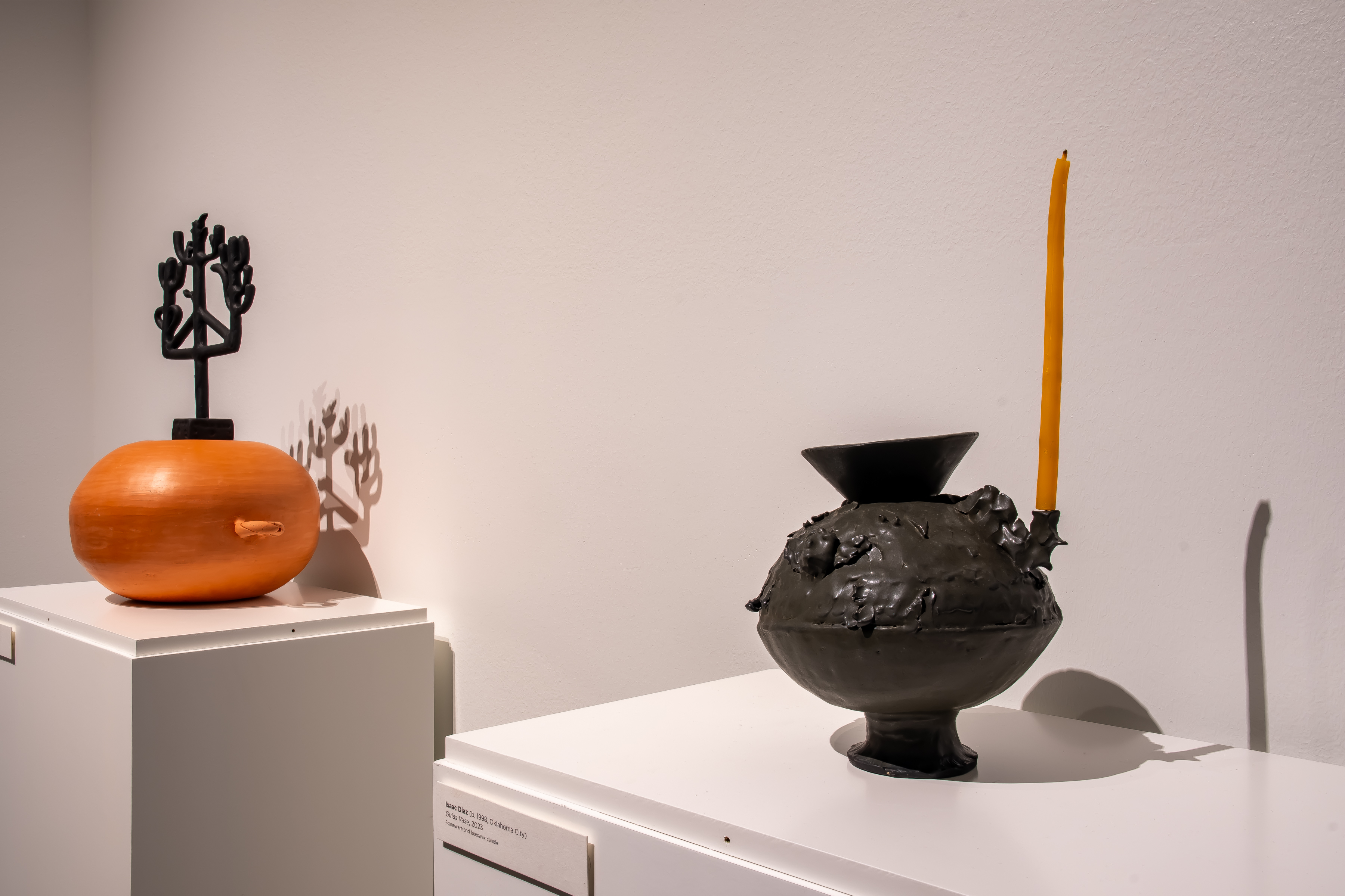Two ceramic vessels sit on white plinths. On the left, the vessel is orange and holds a black candelabra-like piece on top. On the right, the vessel is black and holds a tall yellow candle on the right side.