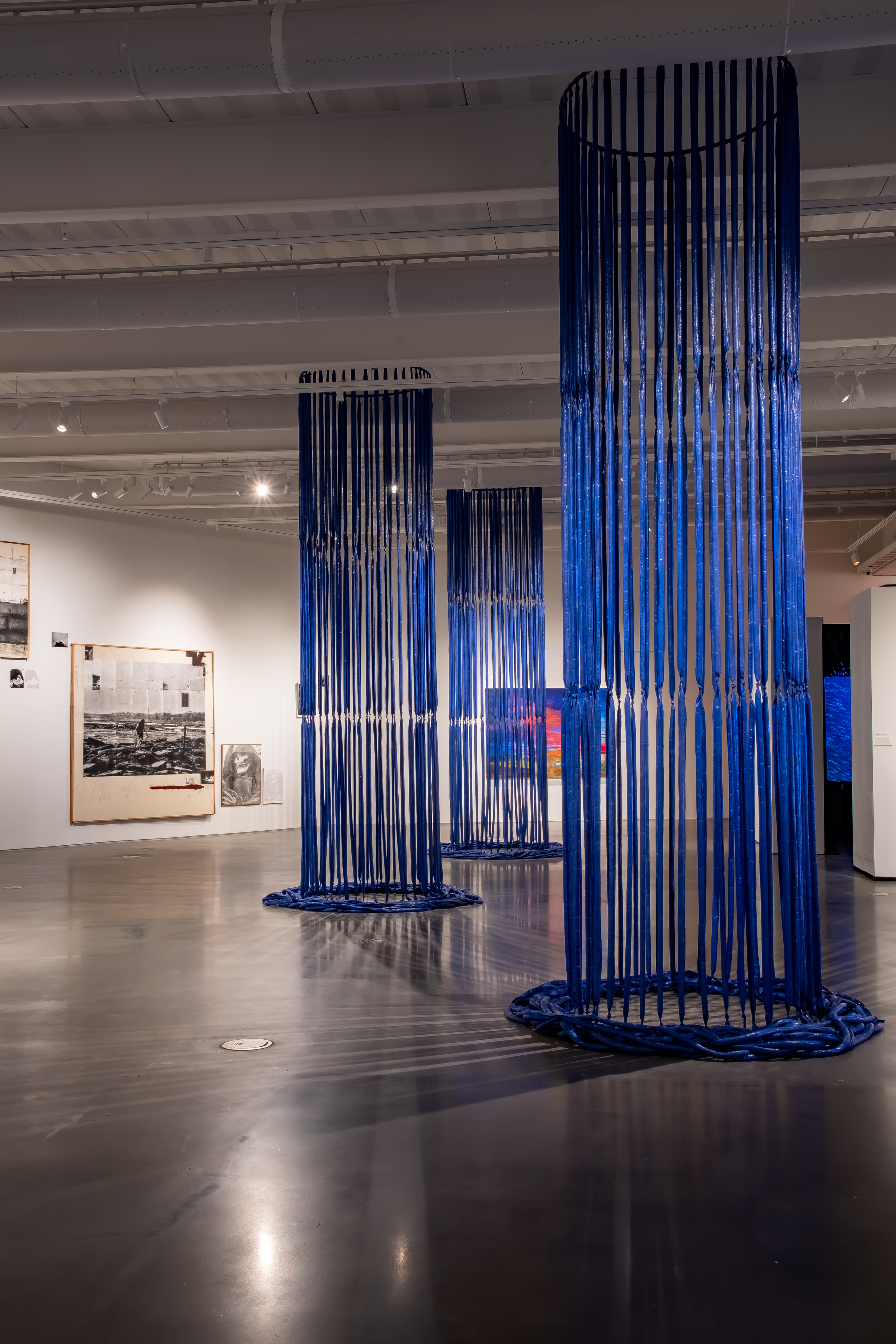 An art gallery. In the center, long flowing columns of blue tarp create cylinders. Three hang staggered behind the other. Black and white photos on a large canvas hand on the back left wall.