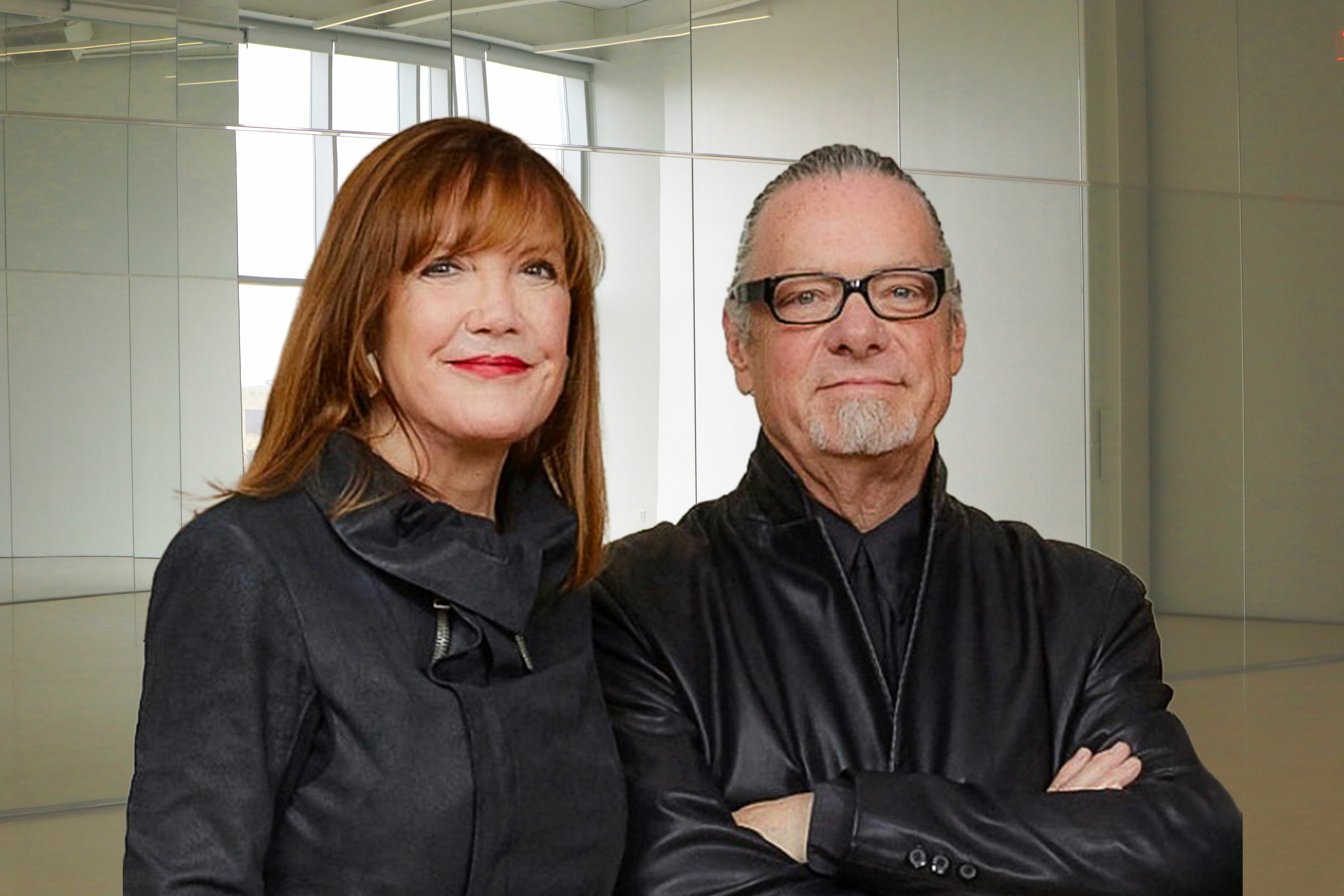 Two older white people stand in a room with mirrors. The woman on the left has red hair, wears red lipstick and is dressed in all black. The man on the right has his hair pulled back and a gray goatee, in a black jacket and wearing black framed glasses.