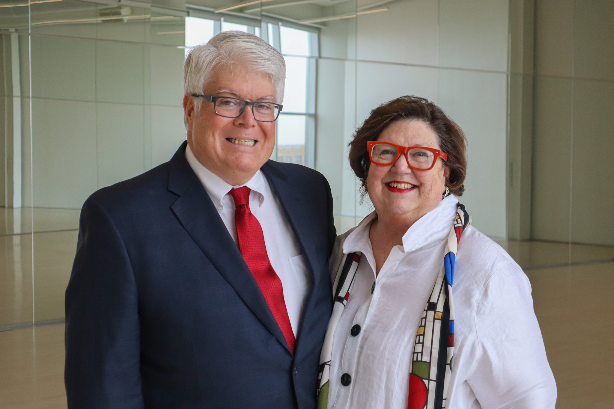 Two older white people stand in a room with walls of mirrors. They are smiling at the camera. The man on the left is wearing a blue suit, black glasses and white tie. The woman on the right is wearing a white shirt, colorful scarf and red glasses.