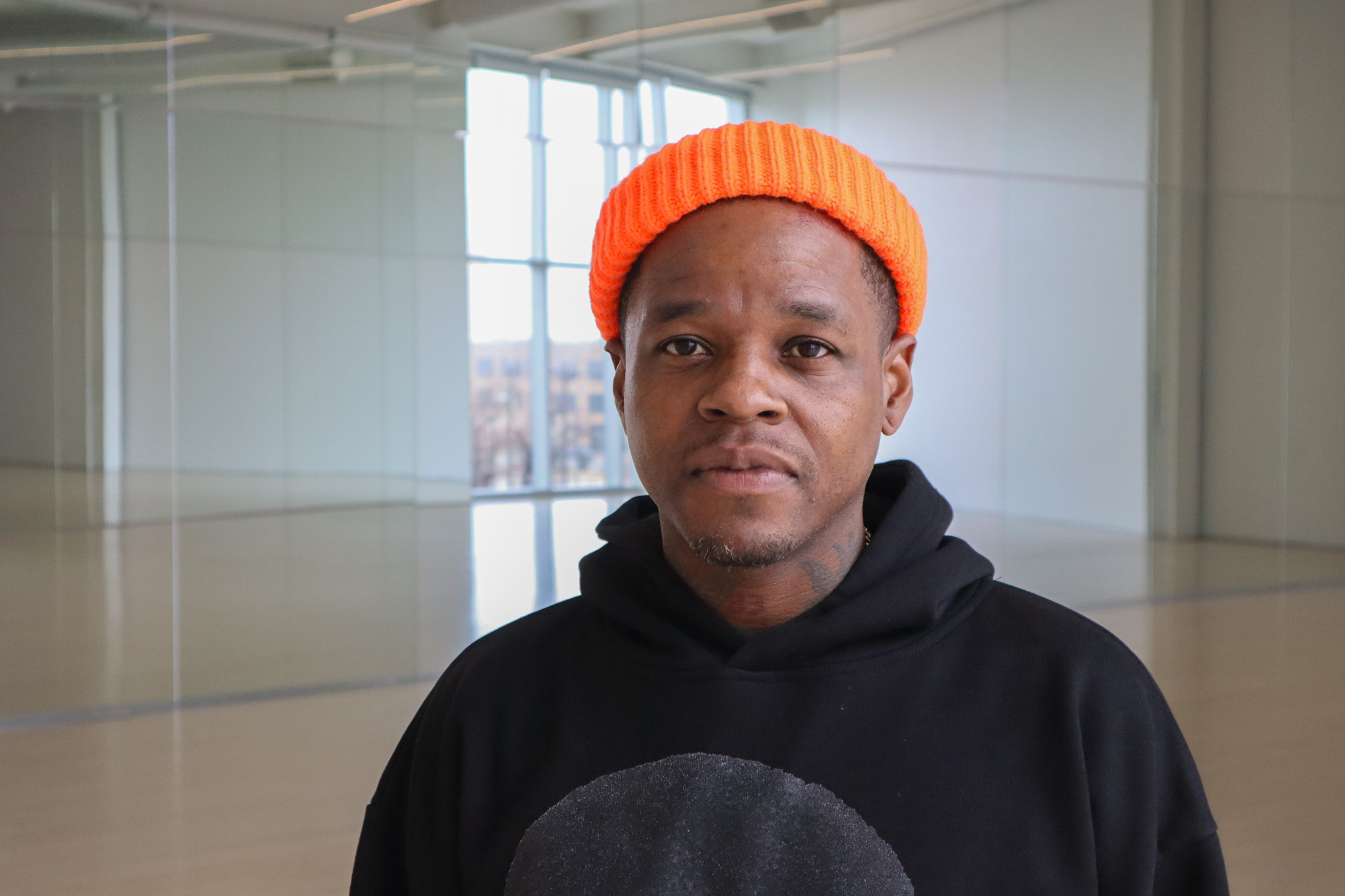 A Black man dressed in a black sweater and wearing an orange beanie stands in a room with mirrors.